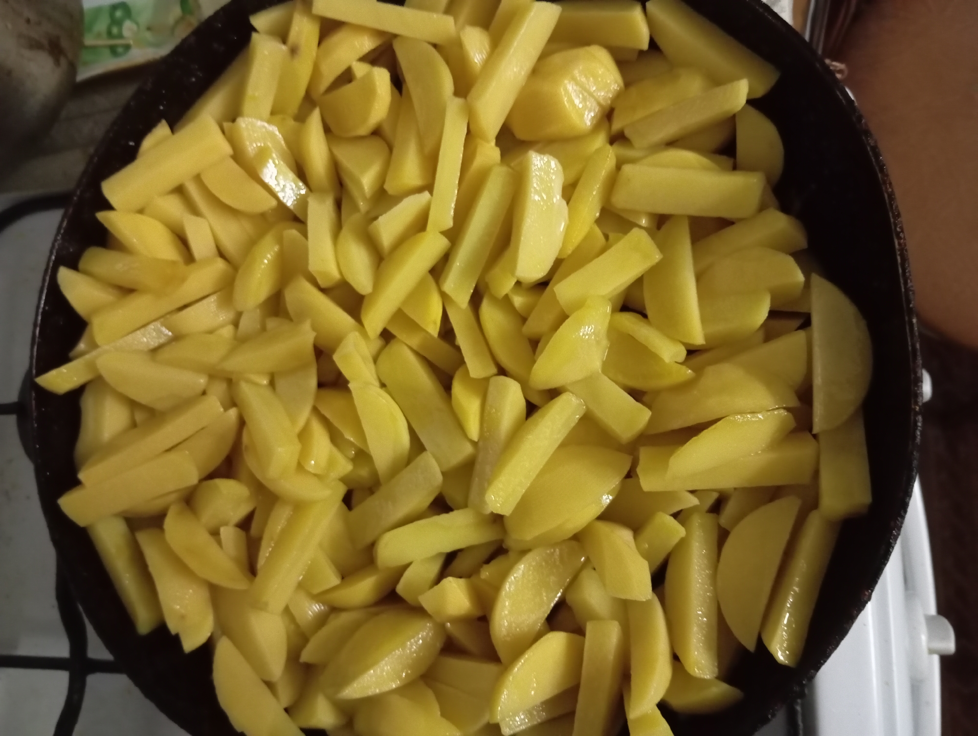 A top-down view of a black frying pan filled with thinly sliced raw potatoes. The potatoes are cut into various shapes, including wedges, batons, and irregular pieces. They appear to be fresh and have a pale yellow, slightly waxy texture, with some pieces reflecting light, suggesting they may be slightly moist. The pan is set on a stovetop, with visible white tiles and black grates of the stove in the background. The overall scene is a close-up shot focusing entirely on the raw potatoes in the pan, indicating preparation for cooking. The location is Zaporizhzhia, Ukraine.