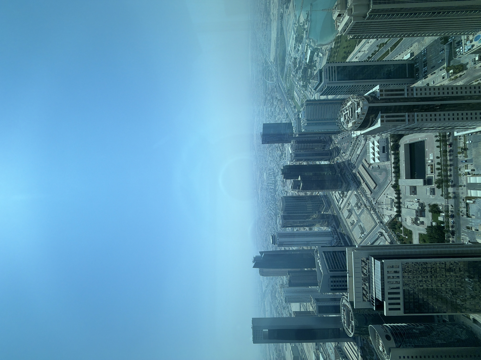 An aerial view captures the modern cityscape of Doha, Qatar, under a hazy blue sky. The perspective is from a high vantage point, looking down onto a dense collection of skyscrapers. The buildings are predominantly glass and steel, with various architectural styles, including sleek, modern towers and some with more intricate facades.
In the foreground, several prominent skyscrapers dominate the view. One building on the far left has a dark, reflective surface and a unique angular top. Adjacent to it are a cluster of narrower, taller buildings with lighter-colored exteriors. To the right of these, a particularly striking building features a rounded, almost curved top, with multiple vertical window sections. Further to the right, another tall, slender skyscraper with a dark, reflective glass exterior stands out. On the far right, a densely ribbed skyscraper forms the edge of the frame.
The middle ground reveals a vast expanse of the city, with numerous other buildings of varying heights and designs spread across the landscape. Roads and infrastructure are visible, including what appears to be a large highway interchange with multiple lanes curving through the urban fabric. In the distance, the city fades into a hazy horizon, with the sky a uniform light blue with a subtle, diffuse rainbow-like phenomenon.
To the right of the city, a body of turquoise water, likely the Arabian Gulf, can be seen. Along the waterfront, some low-lying structures and green spaces are visible. The overall impression is one of a rapidly developed, modern metropolis. No individuals or specific actions are discernible in the image due to the aerial perspective and the distance. The environment is characterized by urban development and a clear, albeit hazy, sky.