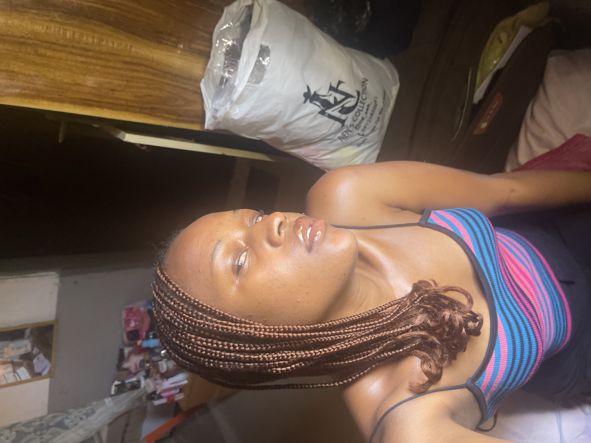 A close-up, low-angle, portrait shot shows a young Black woman with braided hair lying down. She is wearing a colorful striped top with thin straps, featuring pink, blue, and black stripes. Her skin appears to be glistening. Her eyes are open and looking upwards, and her lips are glossed.
In the background, a white plastic bag with black text and a graphic of a cowboy hat and horse is visible, resting on a wooden surface. Other objects and a wall are also discernible. The lighting suggests it might be indoors during the daytime. The overall mood appears relaxed. The visible text on the bag reads: "MEN'S COLLECTION."