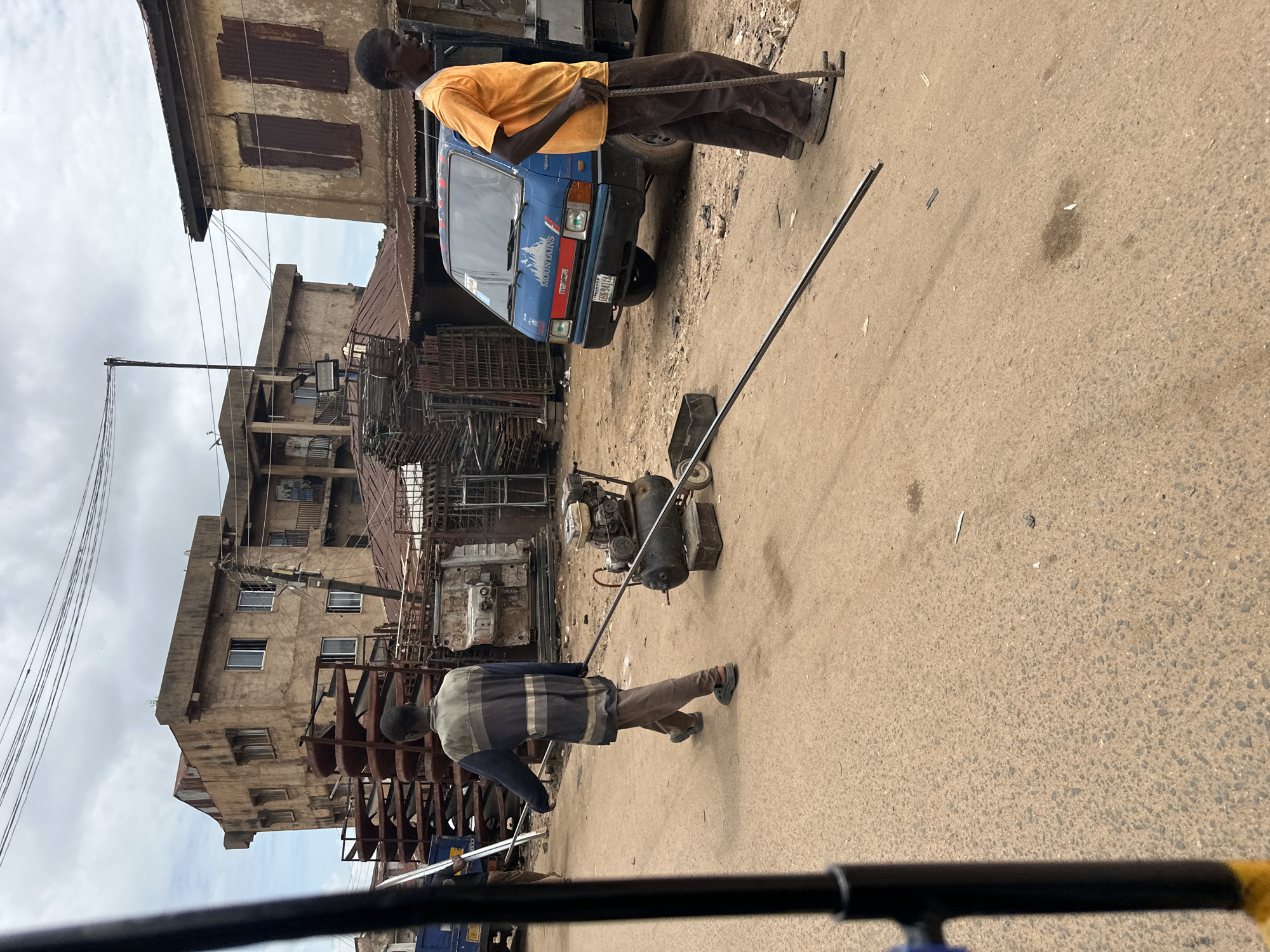 The image depicts an outdoor scene in Jos, Nigeria, characterized by a dusty, unpaved road. In the foreground, two men are engaged in activity. One man, wearing a dark blue and grey striped jacket and brown trousers, is walking away from the camera, carrying a long metal rod. He is also pulling a portable air compressor with a metal frame on wheels. The other man, wearing an orange t-shirt and brown trousers, stands facing towards the right, holding a metal rod with both hands and resting it on the ground. Behind them, a blue Mazda truck is parked. The background reveals a densely built environment with older, somewhat dilapidated buildings made of concrete and corrugated iron roofing. Stacks of metal shelving units and other scrap metal materials are visible, suggesting a commercial or industrial area, possibly a scrapyard or metal fabrication workshop. The sky is overcast, indicating a cloudy day. The overall impression is one of a working street scene in an urban setting.