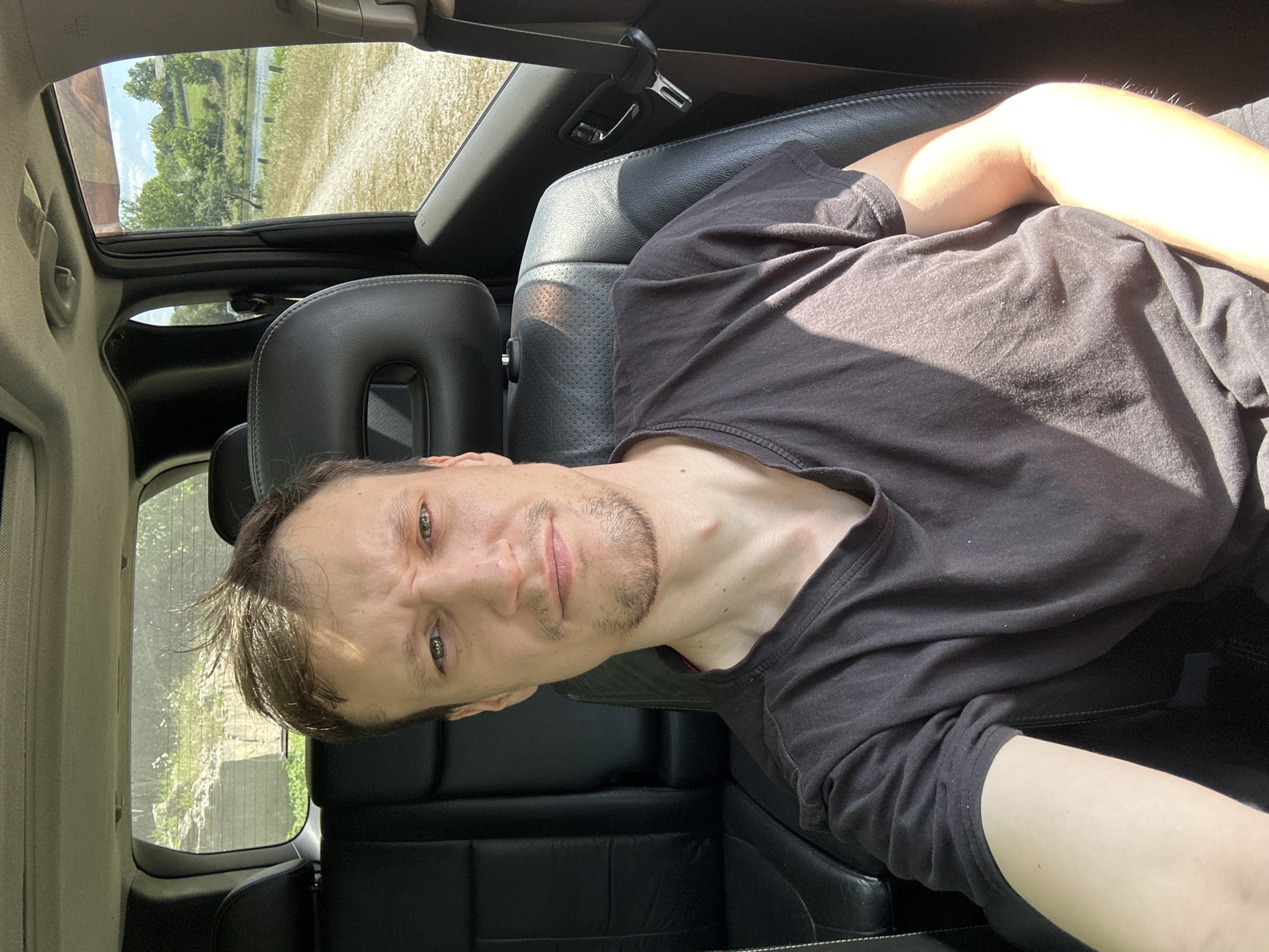 A medium shot shows a man sitting in the driver's seat of a dark-colored car. He is wearing a black t-shirt and his hair is tousled by the wind. The car's interior is visible, with black leather seats and the back seat. Outside the car, a rural landscape is visible, with a dirt road, a body of water, trees, and green hills. The sun is shining, casting shadows on the man's face and the car's interior. The image appears to have been taken in Makiv, Ukraine.