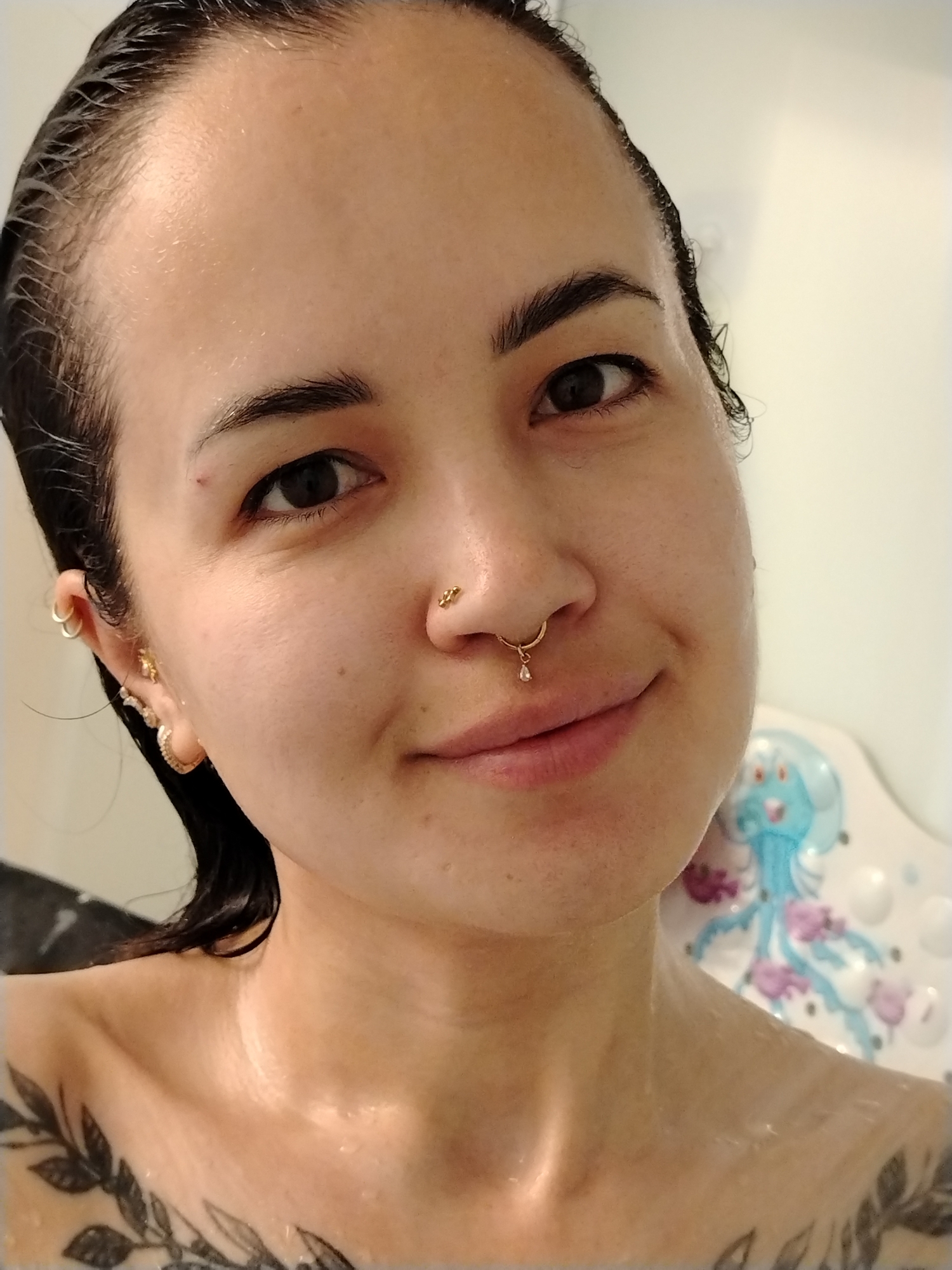 A close-up selfie of a woman with wet, dark hair pulled back from her face. She has light skin, dark eyes, and arched eyebrows. She wears multiple gold earrings in her left ear and gold jewelry in her nose, including a stud and a septum ring with a dangling gem. A floral tattoo is visible on her left shoulder and chest. She is likely in a bathroom or shower, as water droplets are visible on her skin and a white surface with a blue and purple jellyfish design is visible behind her right shoulder. Noviy, Russia, is not relevant to the content of the image.