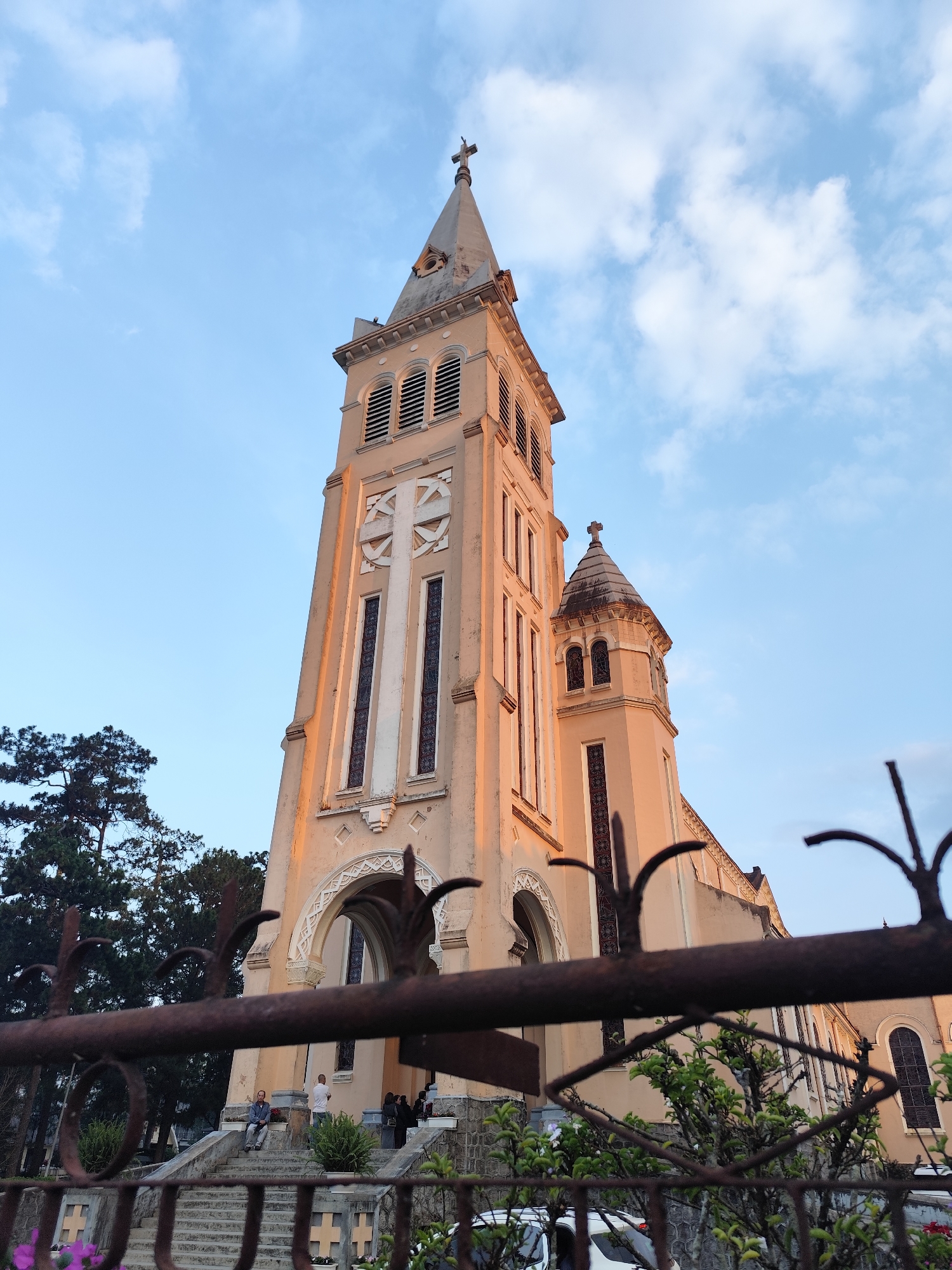The image captures the Saint Nicholas Cathedral (Da Lat Cathedral) in Da Lat, Vietnam, viewed from a low angle behind a wrought-iron fence. The cathedral's bell tower dominates the frame, displaying a light peach-colored facade with a prominent white cross on its front. The sky is a mix of blue and white clouds, suggesting either early morning or late afternoon light. The bottom portion of the image shows a section of a rusty, decorative fence in the foreground. Beyond the fence are steps leading up to the cathedral entrance. Several people are on or near the steps. Some trees are visible on the left side, partially obscuring the lower portions of the cathedral. A white vehicle can be partially seen behind some bushes at the bottom.