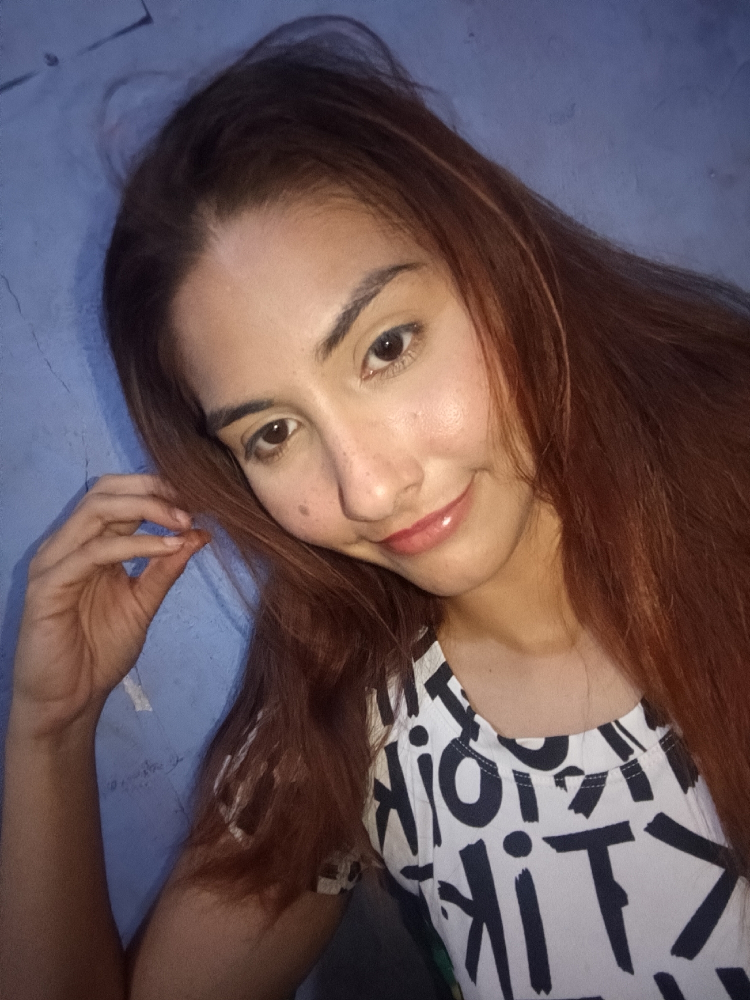 A young woman poses for a selfie. She has light brown skin, dark brown hair, and brown eyes. She is wearing a white tank top with black lettering. Her hand is resting near her head. The background is a light blue wall. The location of Juan Griego, Venezuela, cannot be determined from the image.