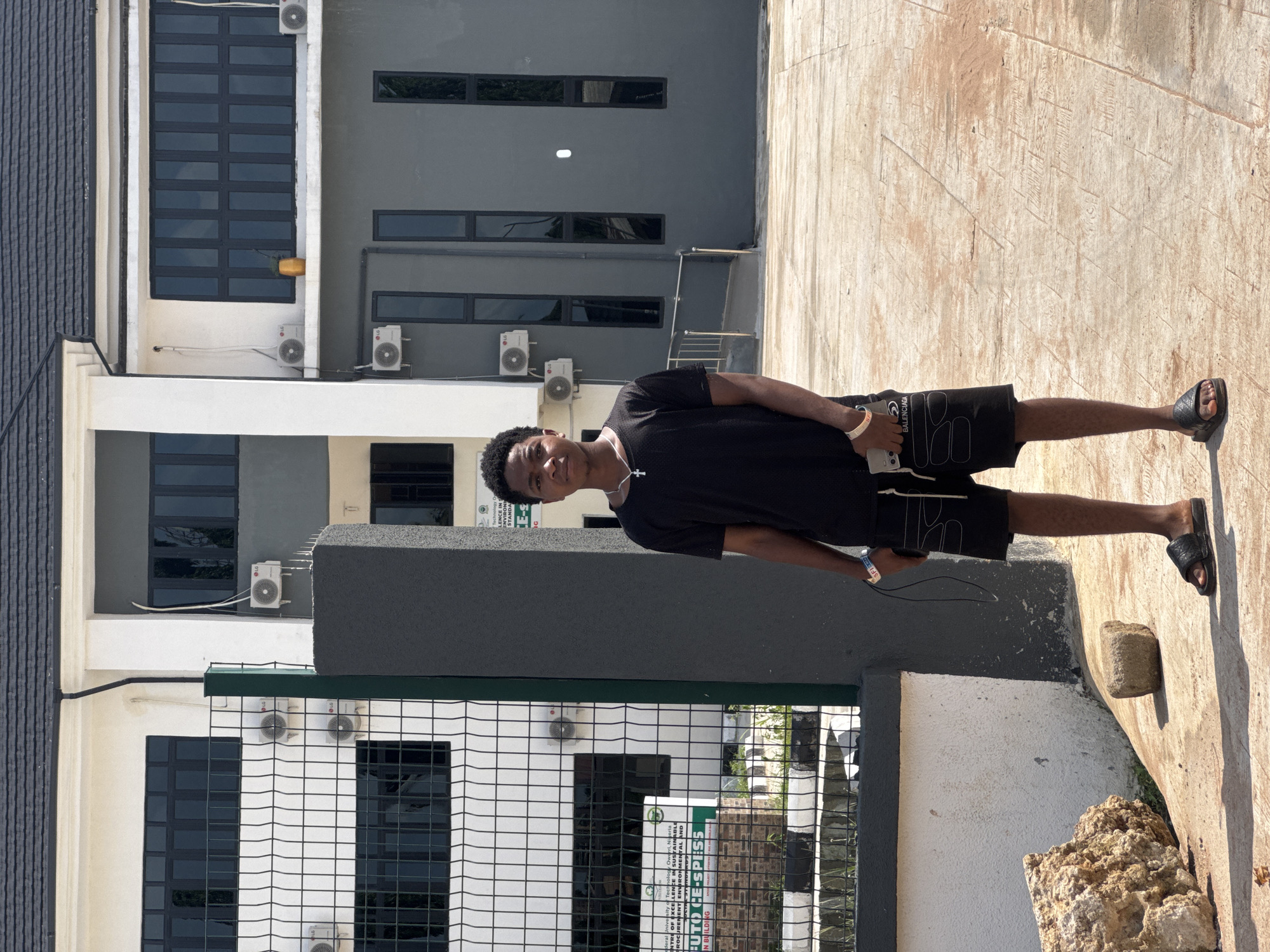 A young man stands outdoors in bright daylight, looking directly at the camera with a slight smile. He is wearing a black t-shirt, black shorts with "BALENCIAGA" visible on the left leg, and black slide sandals. A silver cross necklace hangs around his neck, and a white wristband is on his right wrist. He holds a white smartphone with black earphones plugged in.
The setting is a paved outdoor area next to modern, multi-story buildings, consistent with a campus or office complex in Ihiagwa, Nigeria. The buildings feature grey and white facades with numerous dark-framed windows. Several LG air conditioning units are mounted on the exterior walls at various levels. A dark metal fence or gate is visible in the foreground, separating the man from another part of the building or a different area. The ground is a light-colored pavement, and some rocks or debris are scattered near the man's feet. The strong sunlight casts distinct shadows, indicating a sunny day.
Visible text on signs attached to the buildings includes: "CENTRE OF EXCELLENCE IN SUSTAINABLE DEVELOPMENT, ENVIRONMENTAL AND RESOURCE MANAGEMENT. A CONTRIBUTION TO CE-SPISS MAIN BUILDING", "Ihiagwa, Ni", and parts of phrases like "SFI", "STANDARDS", and "LENCE INS". The brand "LG" is clearly visible on the air conditioning units. The shorts worn by the man have "BALENCIAGA" printed on them.
