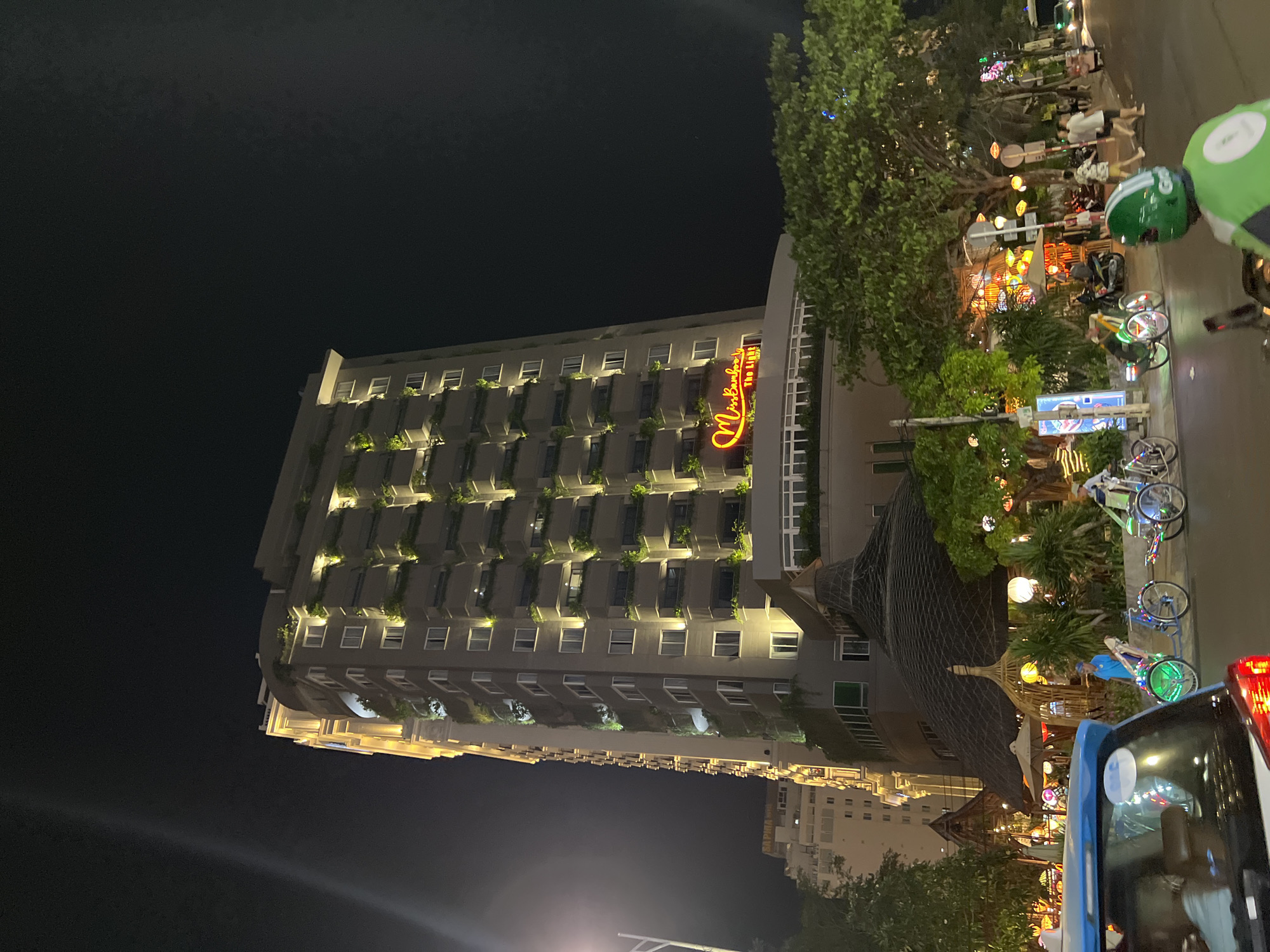 The image depicts a night scene in Nha Trang, Vietnam. Dominating the frame is a tall building with many lit windows and greenery on its balconies. The words "Miss Bamboo" are illuminated in orange signage on the building's side. Adjacent to the building is a street with various lights, trees, and people walking. Bicycles are parked along the sidewalk. The lights, and the people suggest an active nightlife or entertainment area. A vehicle with a red tail light is partially visible in the foreground, and a person wearing a green helmet. The environment is dark, illuminated by artificial light.
