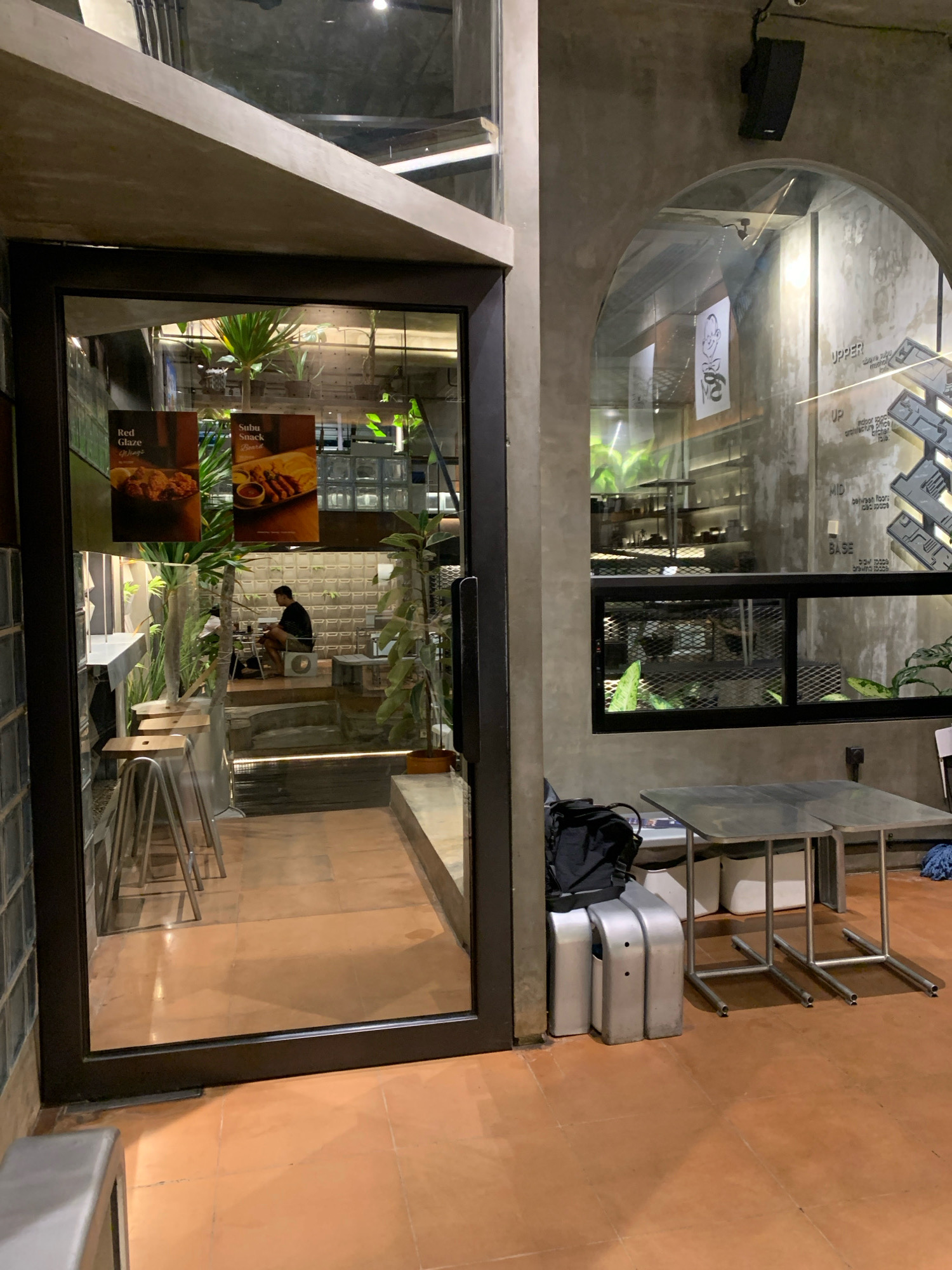 The image shows the interior of a modern cafe with an industrial aesthetic, likely located in Mojokerto, Indonesia. The entrance is a large glass door, revealing a stepped interior with a minimalist design. The walls are exposed concrete, and the flooring is terracotta tile.
Inside, there are a few patrons. One individual, wearing a dark shirt and shorts, is seated at a table on a lower level, appearing to be engaged with their phone. Other seating areas are visible, including high stools with metal legs and wooden tops near the entrance, and smaller metal tables and chairs on the right side of the image.
Decorations include framed pictures of food items on the glass door, such as "Red Glaze Wings" and "Subu Snack Board." Lush green plants are strategically placed throughout the space, adding a touch of nature. The overall ambiance is dimly lit and contemporary, with a focus on clean lines and raw materials. Outside the cafe, on the right, a metal table is set up, suggesting an outdoor seating area or a space for staff. The concrete wall to the right features some graffiti or artwork with text that includes "UPPER," "MID," and "BASE," hinting at different functional zones within the establishment.