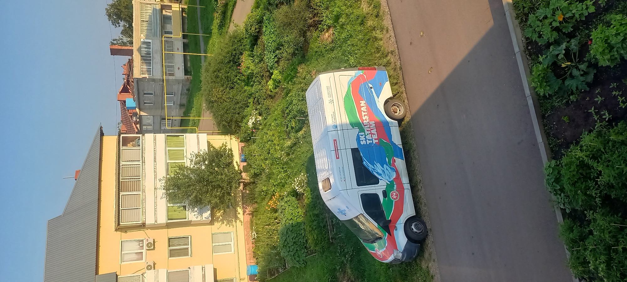 In Menzelinsk, Russia, a white van with "SKI TATARSTAN TEAM" branding is parked on the side of a road adjacent to a patch of green vegetation and a residential building. The van features a colorful design with blue, green, and red wavy lines and a stylized eagle. A garden area with various plants and flowers is visible behind the van, bordering the building. The building itself is a multi-story structure with yellow and cream-colored walls, numerous windows, and balconies. The scene appears to be set during daylight hours with clear skies.