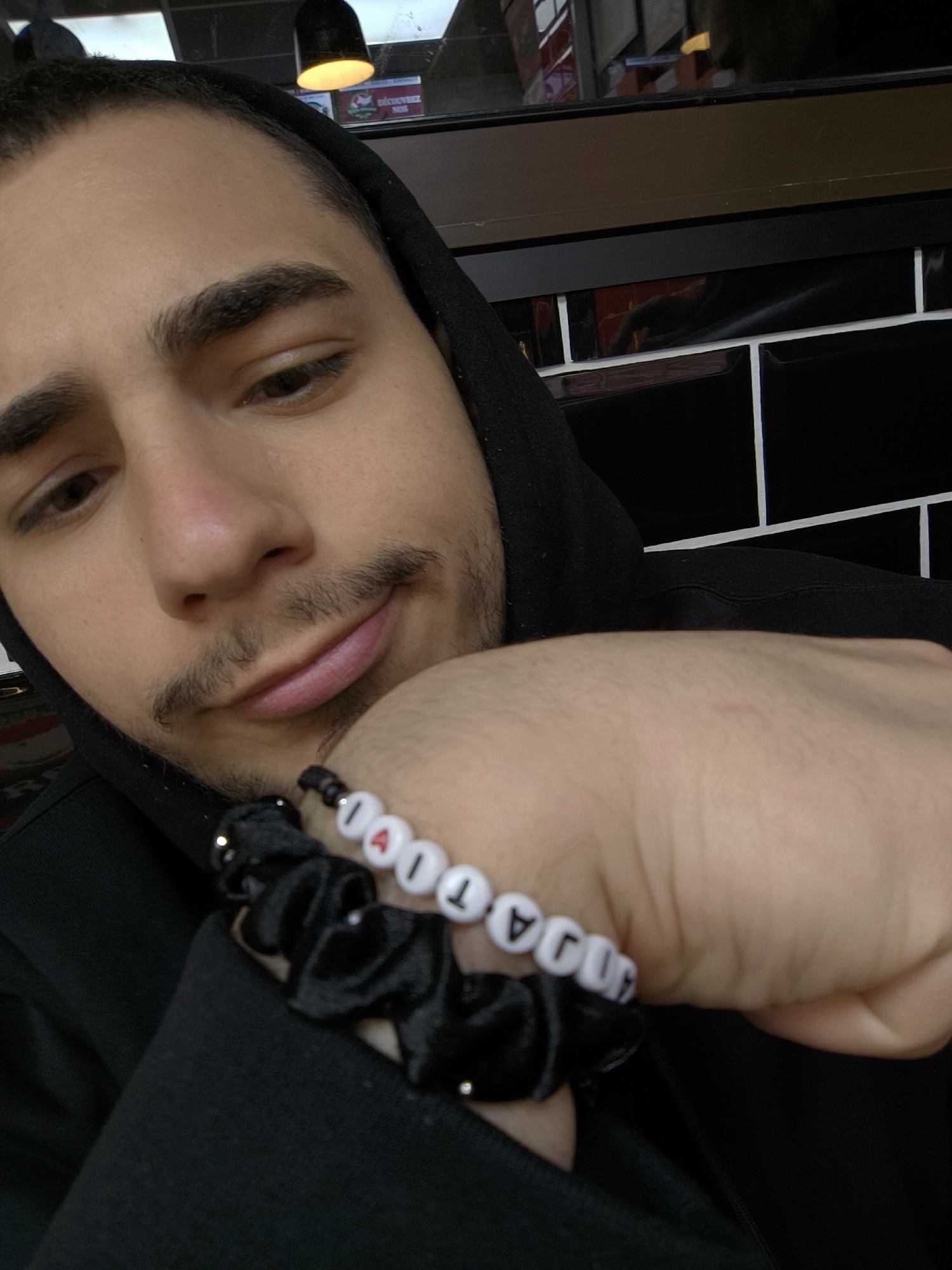 A selfie shot shows a man wearing a black hoodie. He is fair-skinned with dark hair and a mustache. He is looking down, focusing on his right wrist, which rests against his chest. The man has two bracelets on his wrist. One is a beaded bracelet with white beads and black letters, which spell out a word. One bead is red and shaped like a heart. Below that bracelet is a black scrunchie with small silver beads. The background is a black tiled wall with white grout. Above the wall is a wooden trim. There is a light fixture visible in the top center of the image, and reflections can be seen in the wooden trim. Based on the image's context, it is most likely the man is in a restaurant or cafe in Strasbourg, France.