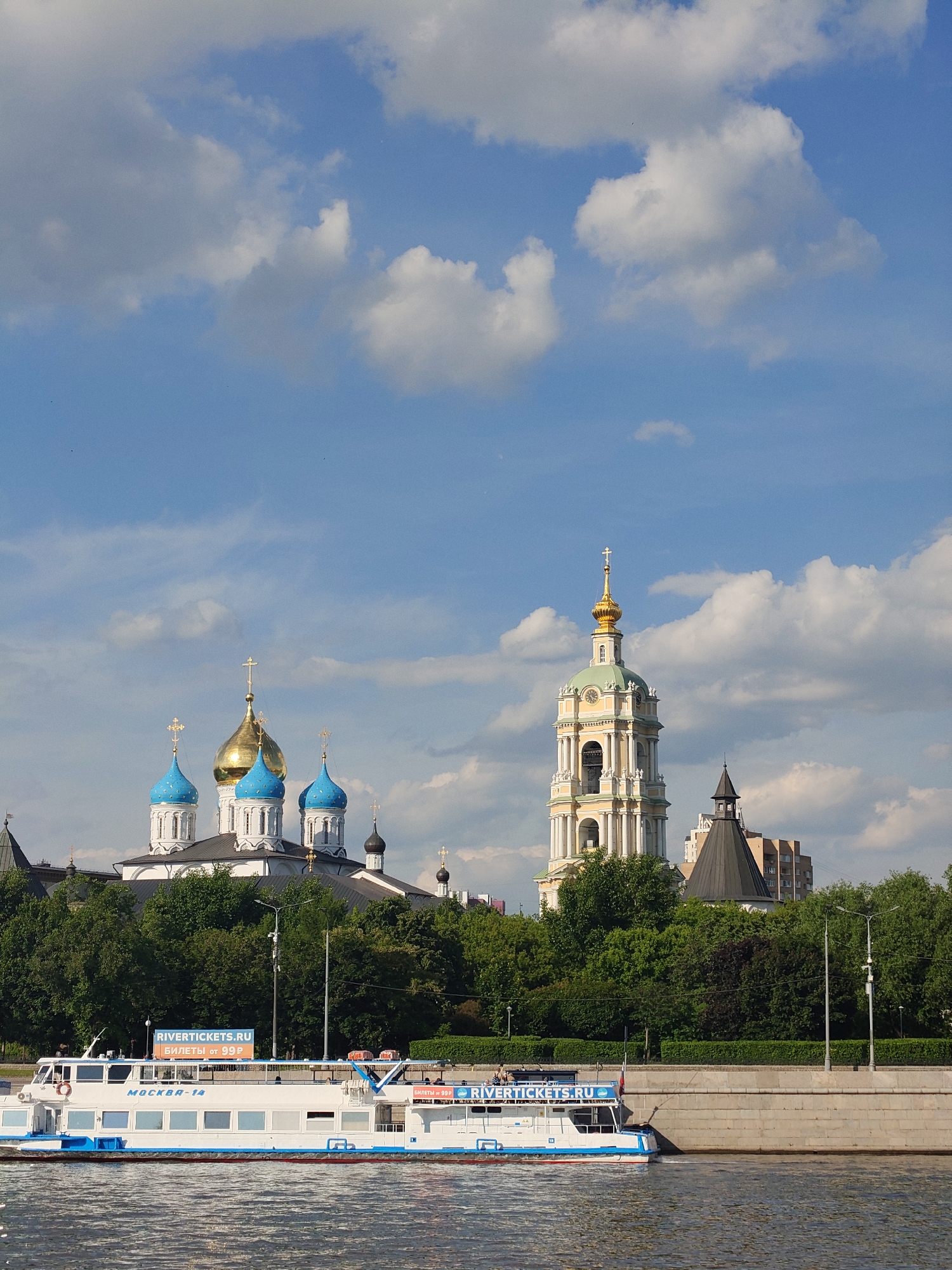 A bright daytime scene captures a large white and blue passenger boat navigating a wide river. In the background, distinct architectural landmarks rise above a dense green tree line along the riverbank.
To the left, a church complex features multiple white buildings crowned with bright blue onion domes and a central shimmering golden dome, all topped with Orthodox crosses. To the right, a tall, elegant bell tower, predominantly light yellow with ornate details, a green dome, and a golden spire, stands prominently, also bearing a cross. Other structures, including parts of modern buildings, are visible between and behind the religious edifices. The sky is a vivid blue with scattered, fluffy white clouds, suggesting clear and sunny weather.
The boat, named "MOCKBA-14" (Moskva-14), has multiple decks and large windows. On its upper deck, a prominent sign displays "RIVERTICKETS.RU" in blue and "БИЛЕТЫ ОТ 99Р" (Tickets from 99R) in orange, indicating it is a tour or transport vessel. People can be seen on the boat's upper deck. The river's surface is gently rippled, reflecting the bright sunlight. A sturdy concrete embankment separates the water from the trees and land beyond. The scene evokes a sense of calm and leisure, typical of a sightseeing tour on a pleasant day. The boat's name and the visible text strongly suggest the location is Moscow, Russia.