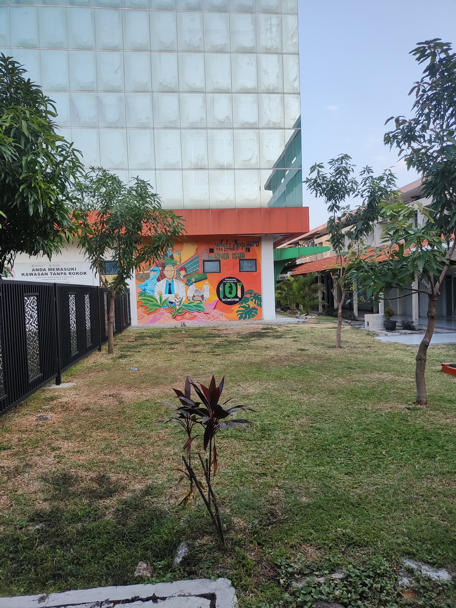 The image captures an outdoor scene in the city of Cirebon, Indonesia, likely on a clear day given the blue sky. In the background, a modern building with a glass facade dominates the upper portion of the frame. Below the glass facade, a bright orange section of the building is visible, adorned with a colorful mural. This mural depicts several figures, possibly medical professionals, engaged in various activities, with some holding medical equipment and others wearing protective gear. The mural also features tropical foliage and a stylized circular emblem with the number "10" inside, suggesting an anniversary or significant event.
In the foreground, a manicured lawn stretches across the scene, interspersed with trees. A black metal fence with intricate patterns runs along the left side of the image, separating the lawn from the building. To the left of the mural, a white sign with Indonesian text is mounted on the wall, indicating a "No Smoking Zone" and referencing local regulations. A small, dark-leaved plant stands in the middle of the lawn, and shadows are cast by the trees, indicating the time of day. The overall impression is one of a well-maintained public space or institutional grounds, possibly a hospital, clinic, or educational facility, given the mural's theme.