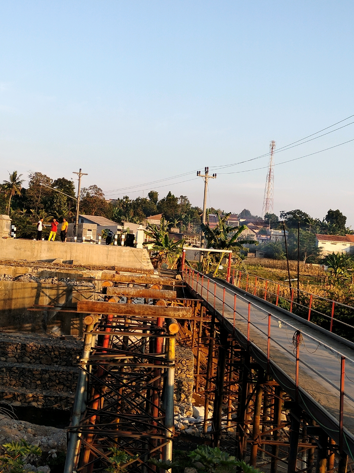 This image shows a construction site in Tersono, Indonesia, where a bridge is being built over a body of water. The bridge is partially constructed, with scaffolding and supports visible. A temporary walkway extends from the land to the bridge. In the background, there are houses, trees, and a radio tower. The sky is clear and blue, with the sun casting long shadows. Three individuals are standing on a concrete embankment, and a motorcycle is parked nearby. The area appears to be rural or semi-rural, with a focus on infrastructure development.