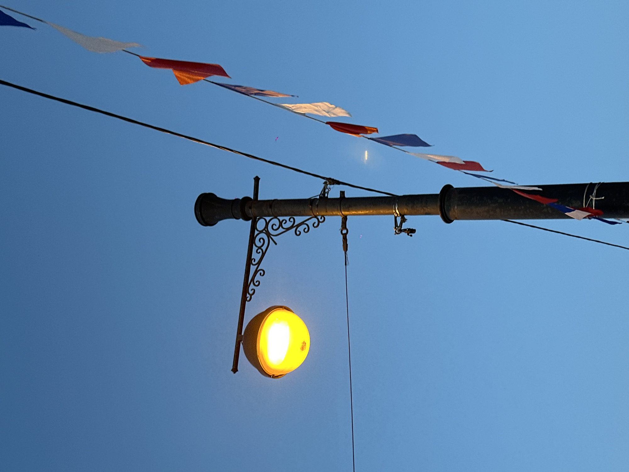 The image shows a street lamp attached to a horizontal pipe with decorative brackets, suspended against a clear blue sky. A string of red, white, and blue pennant flags hangs above the pipe. The location, Irkutsk, Russia, is not discernible from the visual information in the image.