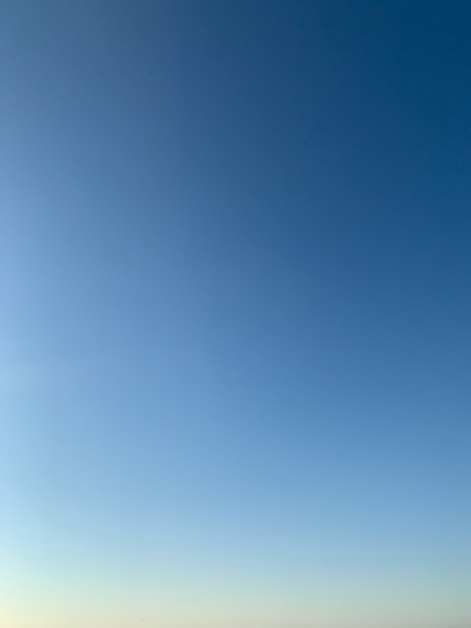 The image shows a clear blue sky with a subtle gradient of lighter blue towards the horizon. The bottom edge of the image shows a faint, pale yellow or white band, suggesting the presence of the horizon or a very distant landmass. No specific landmarks of Comacchio, Italy are visible. The scene is devoid of any people, subjects, or objects. The environment is solely composed of the sky.