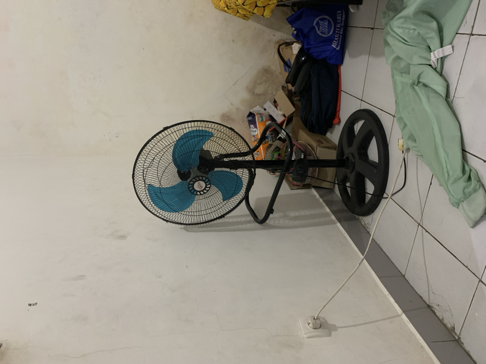 The image captures an indoor scene in Cibinong, Indonesia, depicting a cluttered corner of a room. The primary subject is a standing floor fan with a black metal cage and bright blue blades, positioned prominently on the left side of the frame. The fan's central hub bears partially visible text, possibly "OKAY".
The room's walls are a light, possibly off-white, color and display significant signs of wear and tear, including numerous stains, dirt marks, and visible cracks, particularly near the base and extending across the surfaces. The floor is covered with large, light-colored square tiles that also show grime and some cracks, especially along the edges and grout lines.
In the lower right foreground, a white power outlet is installed on the wall, with a white power cord plugged into it. This cord extends towards the right, disappearing from view, suggesting it powers the fan or another nearby appliance.
To the right of the fan, the corner of the room is filled with various stored items. These include several cardboard boxes, some stacked and appearing either empty or containing miscellaneous goods. Piles of fabric are also visible, most notably a light green sheet or piece of clothing draped on the right, and a patterned yellow fabric near the top edge. A blue bag, possibly a reusable shopping bag or a uniform, is perched atop other items, displaying the white text "BHAKTI KARYA" and "BERSAMA KITA BERKARYA". A dark, circular object, resembling a weight plate, is also discernible amongst the clutter.
No people are present in the image, and no specific activity is taking place. The lighting suggests an interior setting, likely illuminated by ambient daylight or artificial light sources. The overall impression is that of a utility space, storage area, or a general room that is not meticulously maintained.