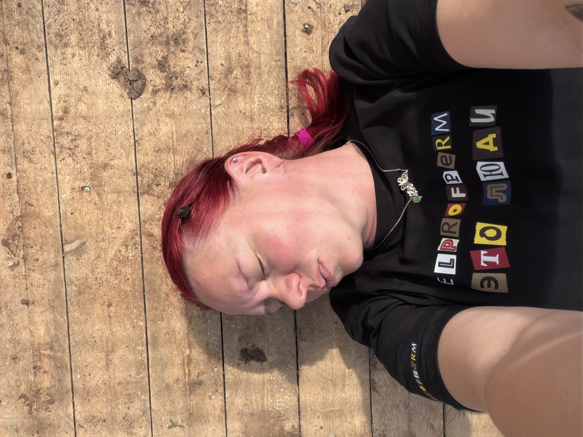 A woman with red hair lies on a wooden surface. She is wearing a black shirt with the Cyrillic text "ELPROFERM это люди". She also has a silver necklace and earrings. The wooden surface appears to be outdoors, possibly a deck or platform. The location may be Chaikovsky, Russia, but there is no way to verify the location from the image.