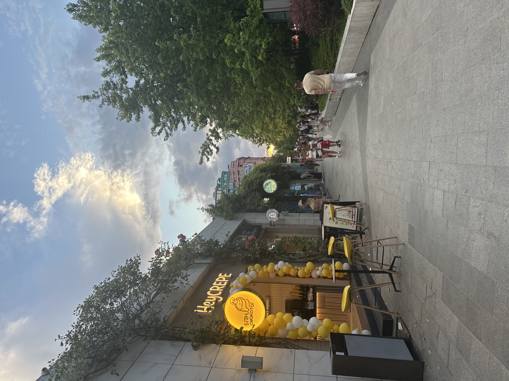 This media file captures a bustling street scene in Odesa, Ukraine, likely during the late afternoon or early evening. The sky is a mix of blue and dramatic, cloudy formations, with hints of sunlight breaking through, suggesting dynamic weather.
The primary focus is on a crepe shop named "Hey Crepe," adorned with a festive garland of white and yellow balloons. The shop's illuminated sign features a cartoonish character and text in what appears to be Cyrillic script. The text includes "Hey Crepe" in English, and below it, "блин, смакота!" and "без черги", which translates to "pancake, delicious!" and "no queue."
In front of the shop, several small tables with yellow chairs are set up, some occupied by customers. A person in a light-colored shirt and white pants is walking away from the camera on the sidewalk. Further down the pedestrian walkway, a line of people, mostly young adults dressed in casual summer attire, are walking or standing, indicating a lively atmosphere.
The setting is an urban environment with buildings visible in the background, some featuring colorful facades. Trees line the walkway, providing shade and greenery. The pavement is made of large, grey stone tiles. The overall mood appears to be energetic and social, with people enjoying the outdoor space and the offerings of the crepe shop.
