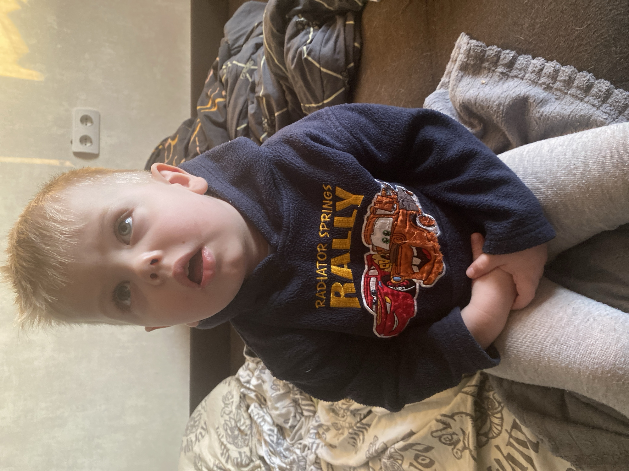A young boy is sitting indoors. He has blond hair and blue eyes. He is wearing a navy blue sweatshirt with a "Cars" themed design and gray sweatpants. He is holding his hands together in his lap. The background includes a bed with various blankets and pillows. An electrical outlet is visible on the wall. The image was taken in the city of Chaikovsky, Russia.