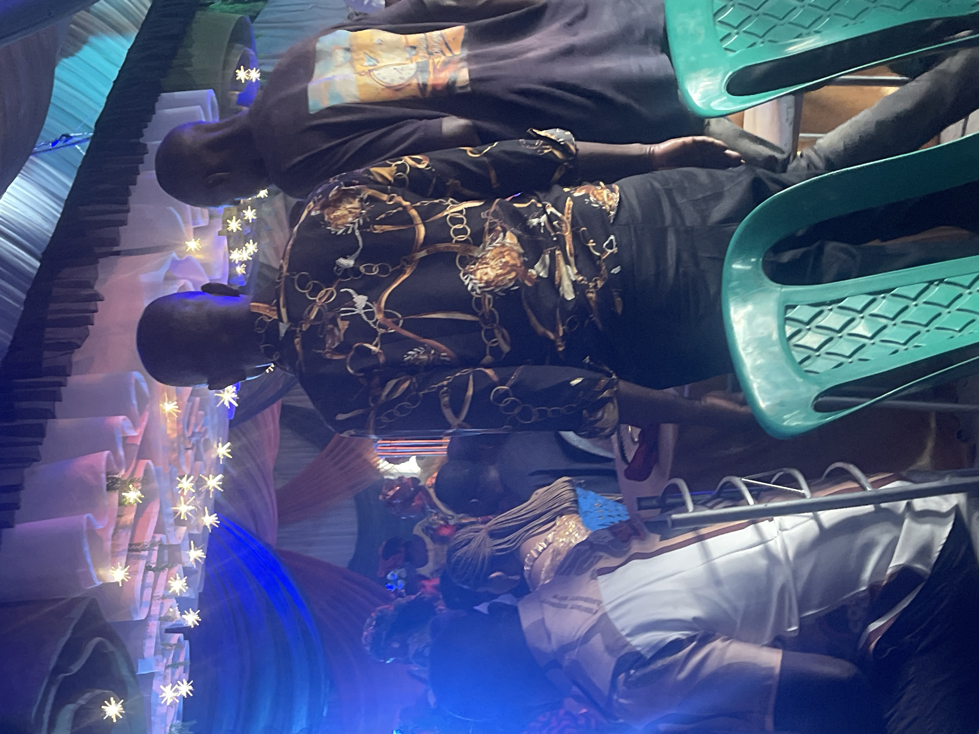 The image shows an indoor setting. Several people are seated on green plastic chairs. A man in a black shirt with a graphic sits in one chair, with another man in a black and gold patterned shirt beside him. In the foreground, a person with braids is wearing a white top. Decorations with lights are visible on the left side. The location is said to be Jos, Nigeria.