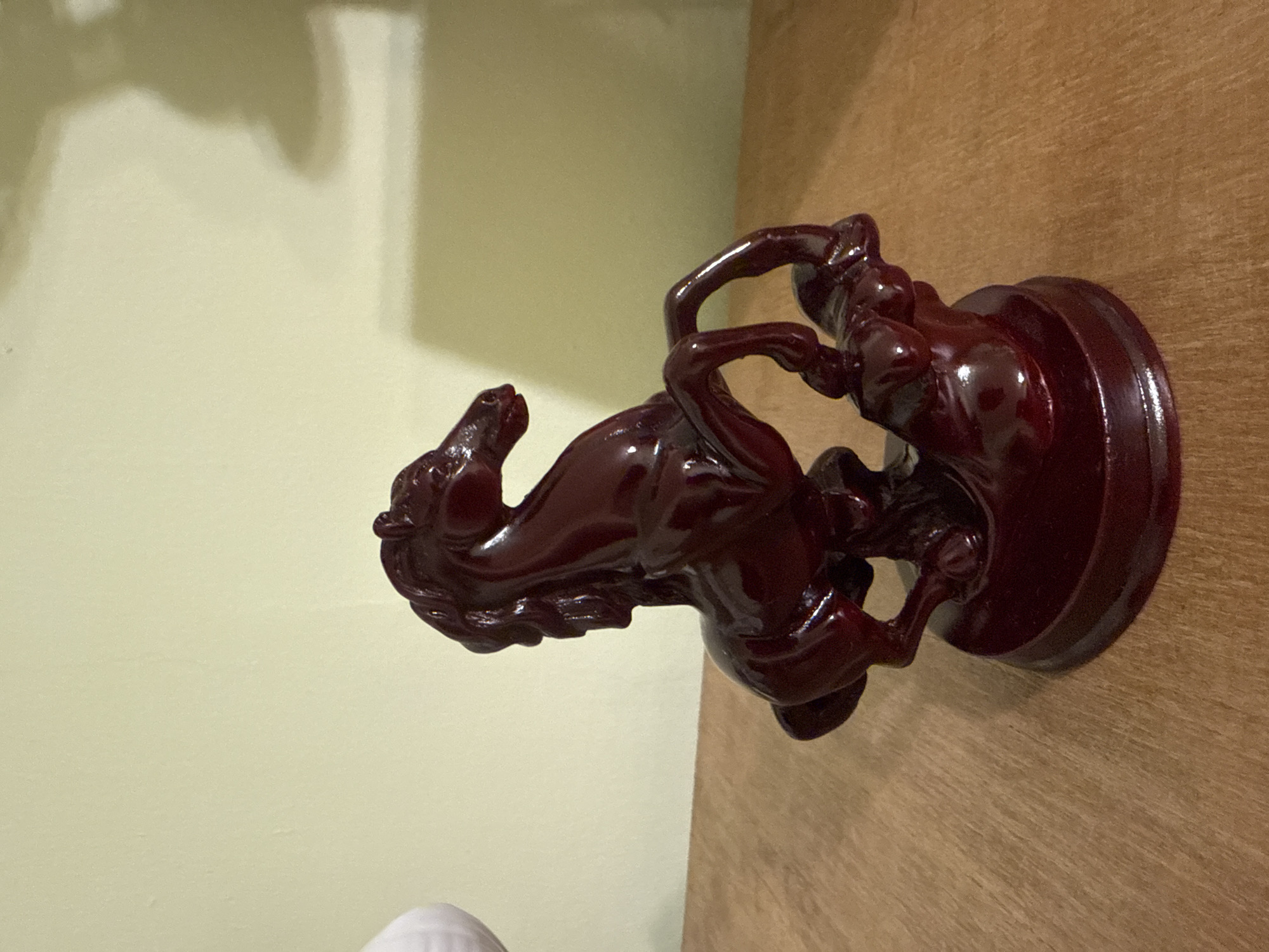 A dark red, high-gloss sculpture of a horse rearing on its hind legs is displayed on a wooden surface. The horse is detailed with a flowing mane and musculature, and it appears to be standing on a rocky base. The background is a plain, light-colored wall, with a soft shadow cast on it. The scene is lit with warm lighting. There is no discernible location in Yerevan, Armenia.