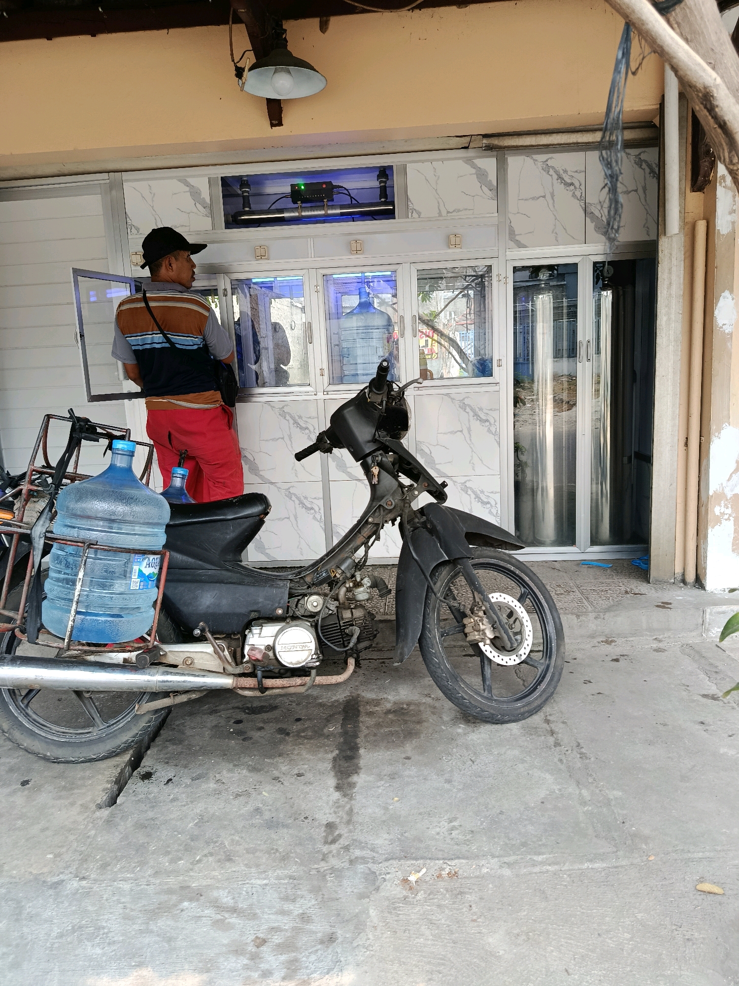 The image depicts a street-level view in Bekasi, Indonesia, featuring a person standing beside a motorcycle. The person, wearing a black cap, a striped shirt, and red pants, is looking towards a storefront. The motorcycle, a black automatic model, is parked in front of the storefront, with two large blue water jugs strapped to its rear rack. The storefront has a white marble-patterned facade with glass doors. Above the storefront, a light fixture is visible. The ground is paved with concrete, and a crack is noticeable in the pavement near the motorcycle's front wheel. The overall setting appears to be an outdoor commercial area in a moderately developed urban environment.