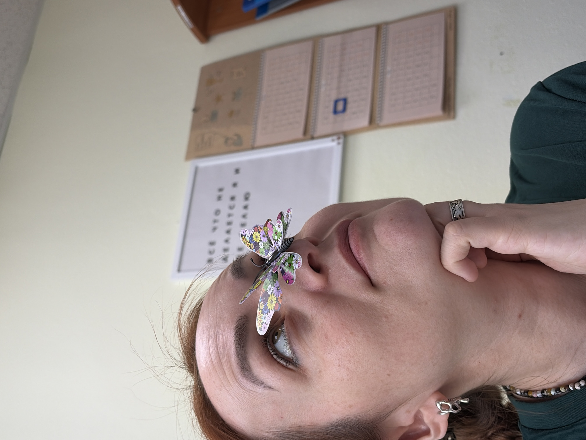 A portrait of a light-skinned person, likely female, with a colorful, paper butterfly resting on their nose. The person has brown hair, fair skin, and is wearing a dark green top. A ring is visible on one of their fingers. The background includes a wall with a whiteboard and what appears to be a calendar hanging above it. Based on the limited information, the location is inconclusive; the reference to Chaikovsky, Russia, cannot be confirmed from the image.