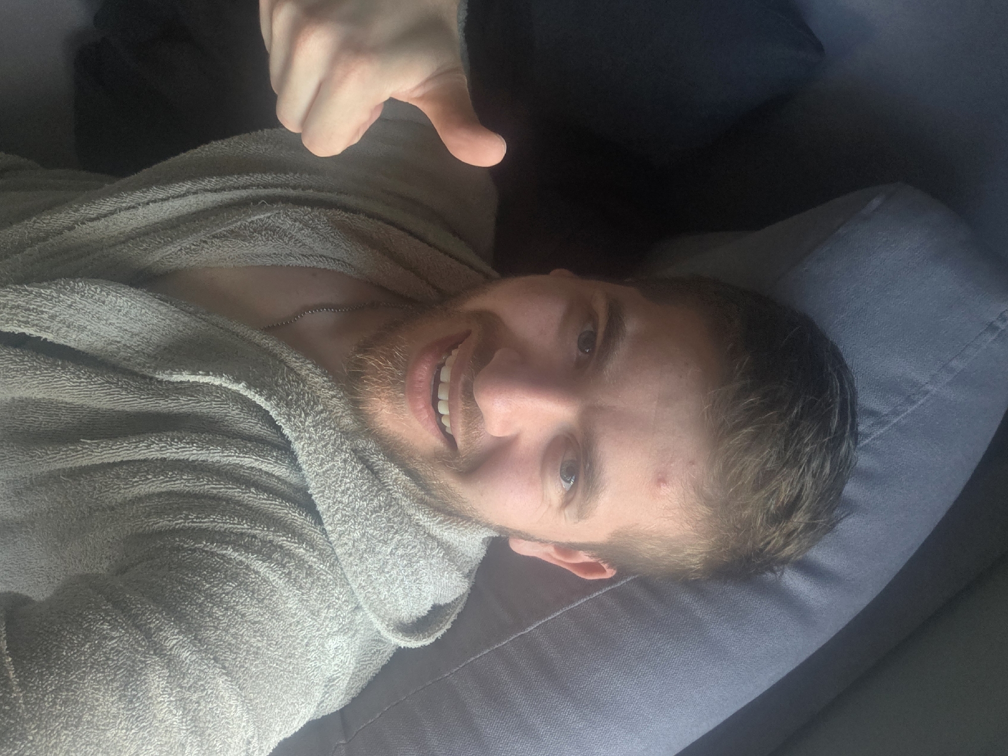 A fair-skinned man with light brown hair and a beard is lying on a grey couch, wearing a grey robe and a thin silver chain necklace. He is smiling and giving a thumbs-up. His head is resting on a grey pillow. The setting is indoors, likely a private residence, but the location is undetermined and there is no way to verify it is Yagodovo, Bulgaria from the image.