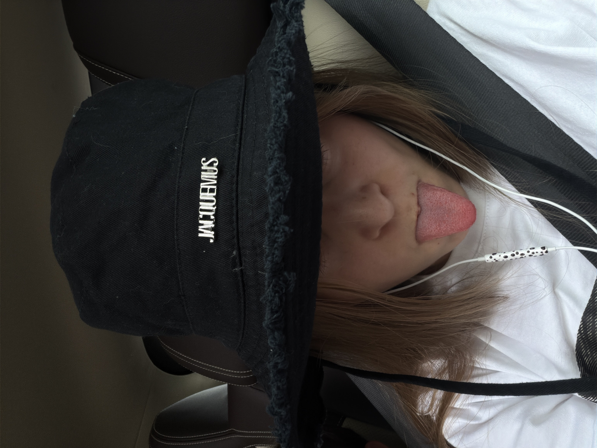A young person, identified as female by their long, light brown hair, is seated in the passenger seat of a vehicle. The person is wearing a black bucket hat with the Jacquemus logo prominently displayed in silver lettering. The hat has a frayed brim. The person is also wearing a white t-shirt and white earbuds with a black and white patterned cord. The person's tongue is sticking out, and their eyes are not fully visible due to the hat. The background suggests the interior of a car, with a dark-colored headrest visible. The scene appears to be within or near the city of Vnukovo, Russia, given the context provided. The overall impression is casual and playful.