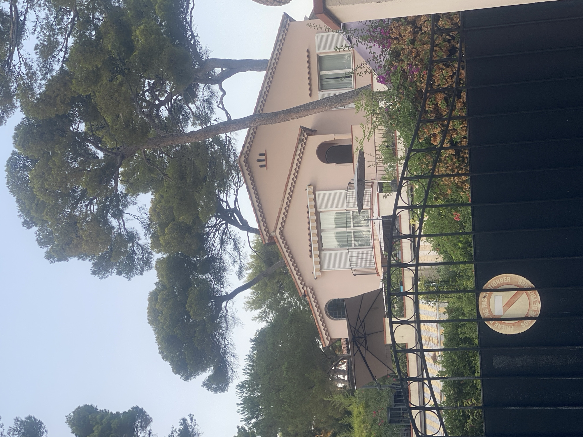 The image shows a pale pink house with a tiled roof, surrounded by tall pine trees. The house has white shutters on its windows and a balcony with a metal railing. In front of the house, there is a black metal gate with a circular sign that reads "INTERDICTION DE STATIONNER SON DE VOITURE" which translates to "NO PARKING PRIVATE PROPERTY". The gate is partially covered by lush green vegetation, including some flowering plants with purple and orange blooms. The background is a clear sky. The scene appears to be a residential area in Antibes, France, with a Mediterranean architectural style and lush vegetation typical of the French Riviera.