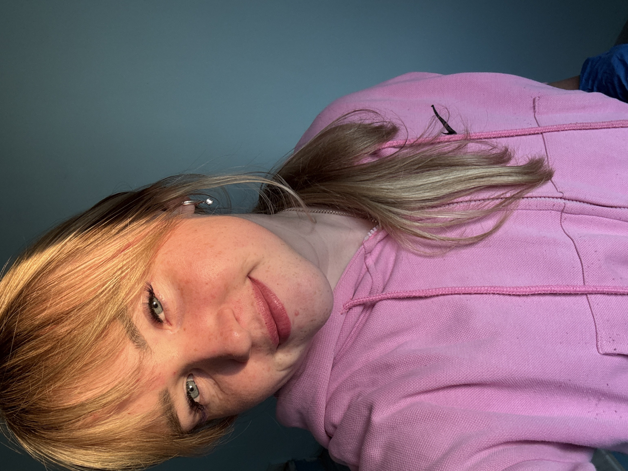 An upward-facing portrait captures a young individual, reportedly in Марковский, Russia, with blonde hair and light-colored eyes, possibly green or blue. The subject is wearing a pink hooded sweatshirt (hoodie) and is looking directly at the camera with a gentle, pleasant smile. Numerous freckles are visible across their nose and cheeks.
The image is rotated 90 degrees counter-clockwise, suggesting the person might be lying down or taking a selfie from a low angle. Blonde hair falls casually around her face, with some strands catching the warm, natural light that illuminates one side of her face and hair. A small, sparkling stud earring is visible in her left earlobe.
The background consists of a plain, solid blue-green or teal wall. The lighting suggests it is daytime, with soft shadows and highlights adding depth to the scene. On the pink hoodie, a dark, elongated logo resembling a Nike swoosh is visible on the upper left side. The fabric of the hoodie shows some texture and minor pilling, and a pink drawstring is evident around the hood opening. A glimpse of another person or object wearing a blue garment can be seen in the far background to the right. The overall mood is casual and serene.