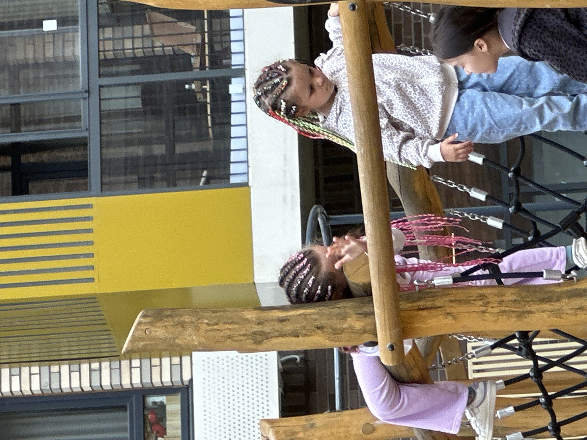 In Znamya Oktyabrya, Russia, a playground scene unfolds. Three young girls are present, engaged with the wooden structures. The first girl, on the left, is leaning against a wooden pole, wearing light purple pants and white sneakers. Her hair is styled in braids. The second girl, in the center, is also interacting with the wooden structure, her hair a mix of braids with colorful accents. She is wearing a light-colored, patterned hoodie and blue jeans. The third girl, on the right, is looking towards the second girl, wearing a dark, patterned jacket. The background reveals a building with yellow and grey vertical accents, windows with metal grating, and a brick facade, suggesting an urban or semi-urban setting. The playground equipment includes wooden beams and black netting with metal connectors.