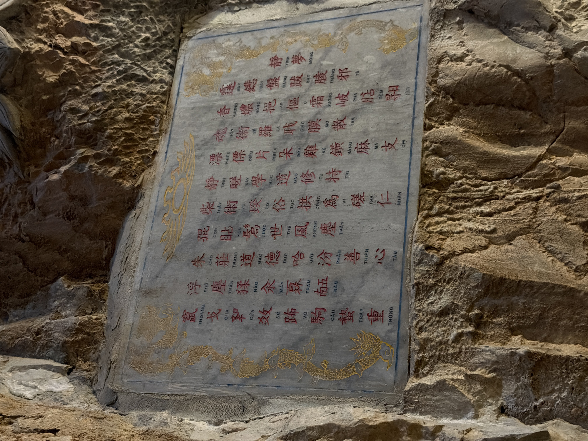 Here's a detailed description of the image:
The image shows a weathered stone tablet embedded in a rock wall, likely within a cave or grotto. The tablet is covered in red and gold calligraphy, featuring traditional Vietnamese text and decorative dragon motifs along the top and bottom edges. The stone of the wall appears rough and aged, with varied textures and earth tones.
While the exact location can't be determined solely from the image, the tablet's style and the rocky cave setting suggest it is potentially located in the Marble Mountains in Đà Nẵng, Vietnam. These mountains are known for their network of caves and Buddhist shrines that often feature similar stone carvings and calligraphy.