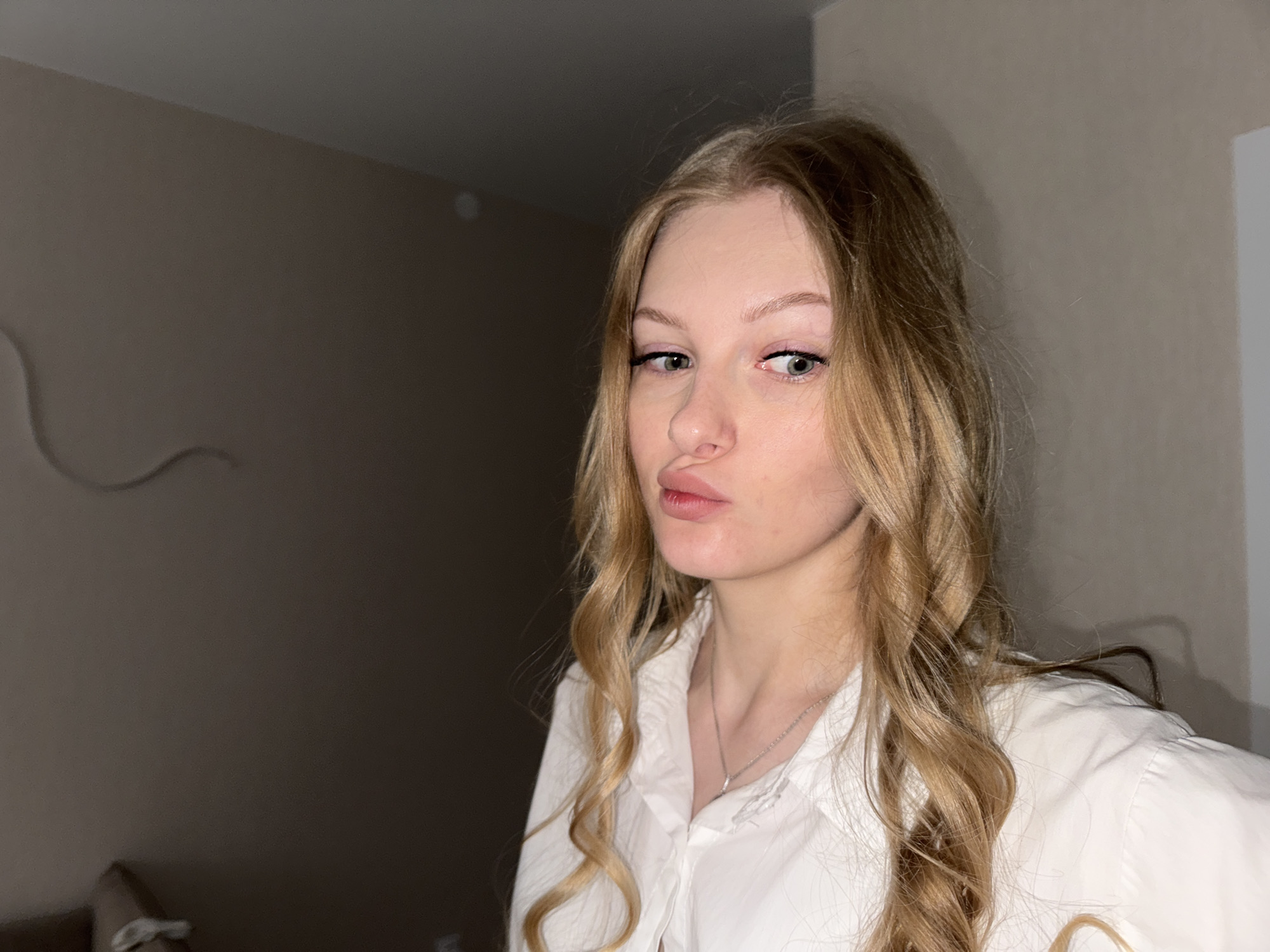 A young woman with long, wavy blonde hair and light-colored eyes is captured in a medium shot, her head tilted slightly to the left. She is wearing a white collared shirt and a thin silver necklace. Her lips are pursed, giving her a playful expression. The background is a plain, muted wall, suggesting an indoor setting. There is no indication of the city of Perm, Russia, or any specific location.