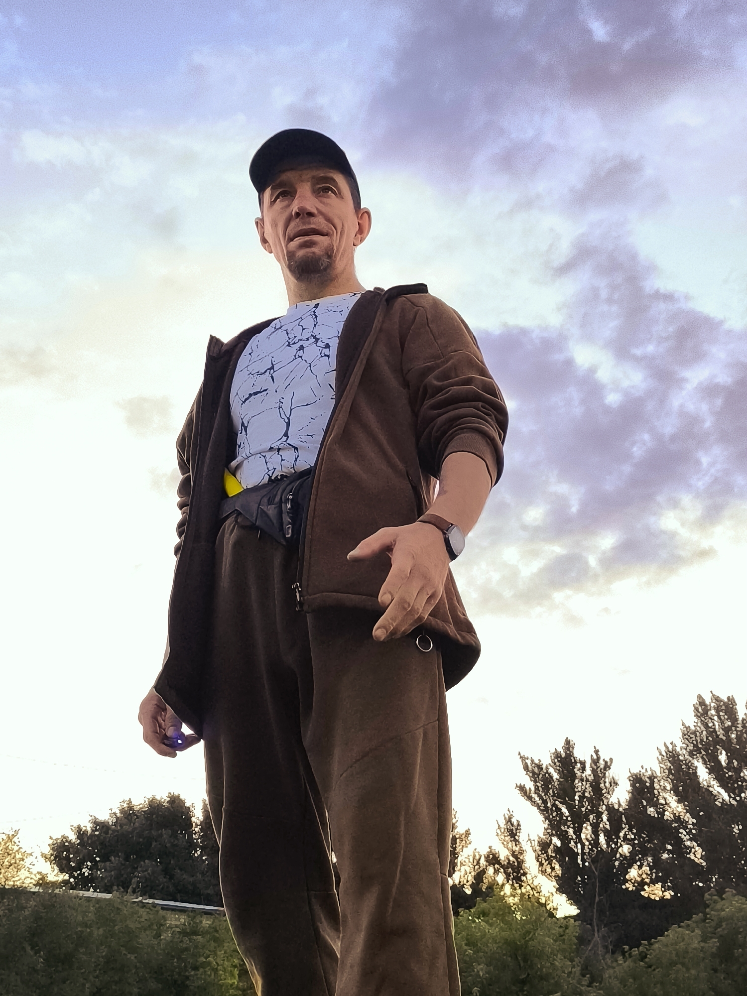 An eye-level, full-body, low-angle shot captures a man standing outdoors with a cloudy sky overhead. The man, wearing a baseball cap, a white t-shirt with a black abstract pattern, and a brown zippered hoodie over brown sweatpants, looks towards the sky. He has a goatee and appears to be in his late 40s or 50s. A black fanny pack is worn around his waist, and a watch is visible on his left wrist. His right hand is raised, and he is holding a small, blue object. In the background, lush green trees and foliage are visible against the bright sky, suggesting a park or natural setting. The image is taken in Qarağandı city administration, Kazakhstan.