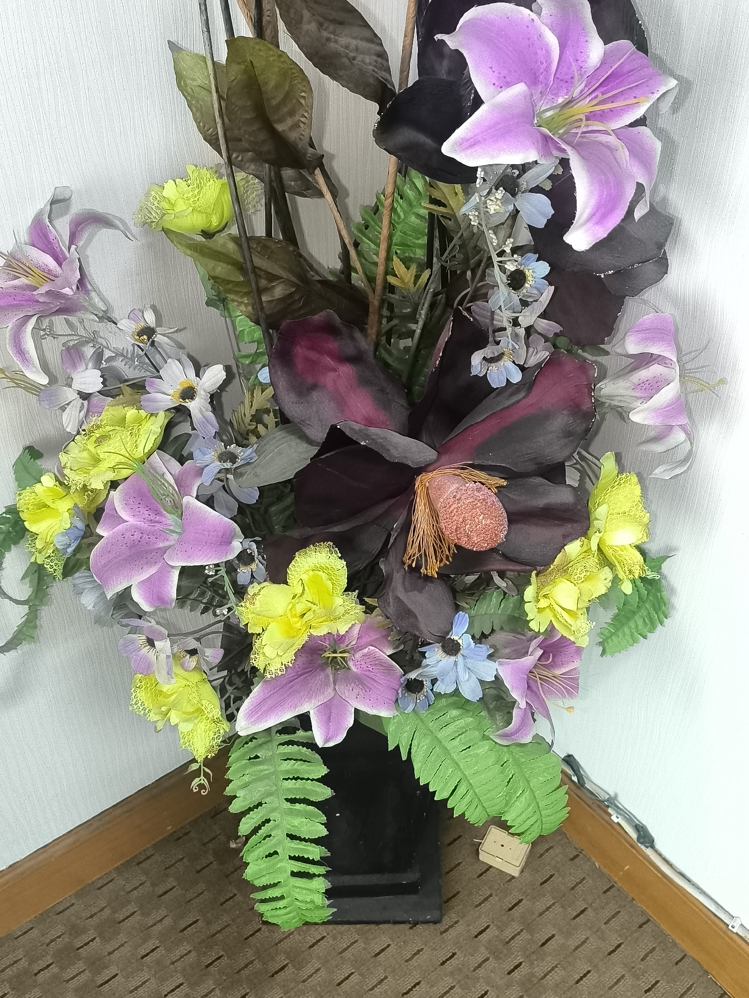 The image shows a large, artificial floral arrangement in a black vase, placed in the corner of a room. The arrangement contains a variety of flowers, including large pink lilies, yellow ruffled flowers, smaller blue flowers, and dark purple/black magnolia-like flowers. There are also artificial green leaves and ferns. The vase is on a brown carpet with a diamond-like pattern. The walls are white and textured. A wooden baseboard runs along the bottom of the walls. A small, light-colored object, possibly an electrical adapter, is on the carpet near the vase. The lighting suggests an indoor environment. The image was taken in Kalabbirang, Indonesia.