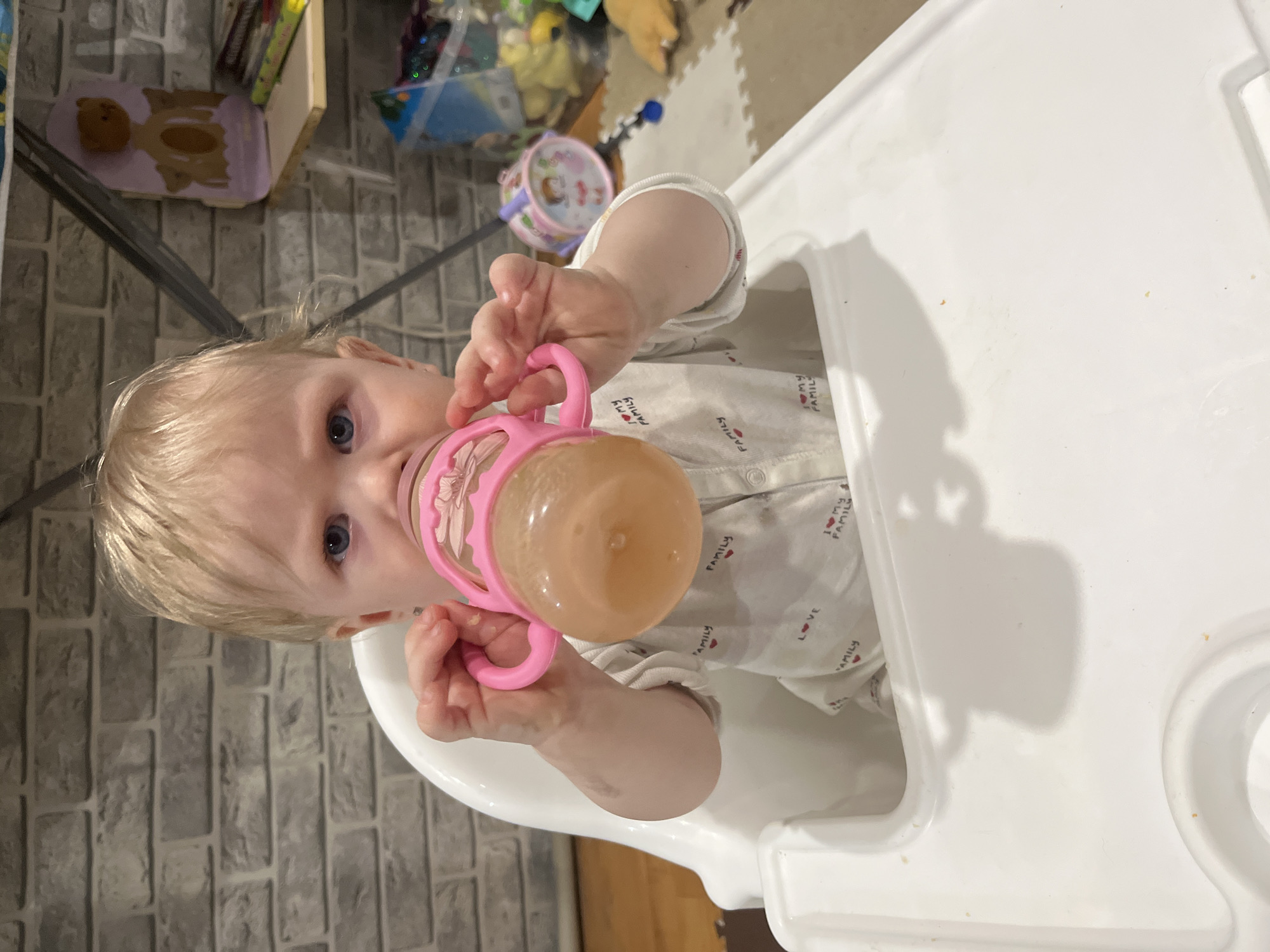 A young child with blonde hair and blue eyes is drinking from a pink sippy cup while sitting in a white high chair. The child is wearing a white shirt with small heart and "Family" prints. The background includes a gray brick wall and various toys and objects on a shelf. The location cannot be confirmed as Vesyoloye, Russia.