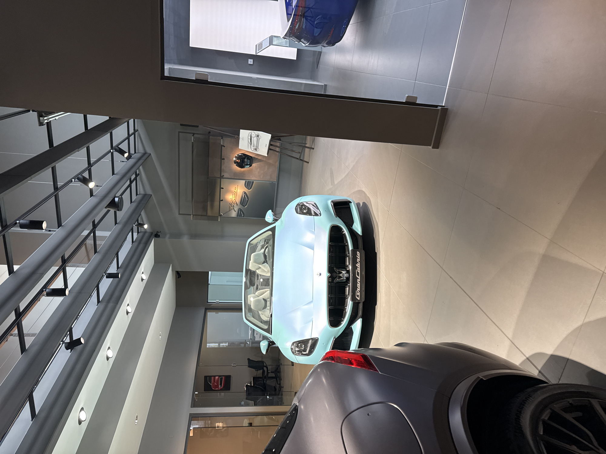 The image shows the interior of what appears to be a car dealership. In the foreground, two cars are partially visible: a blue Maserati GranCabrio and a grey car behind it. The interior has a modern design with grey beams supporting track lighting. Other cars are displayed in the background behind glass partitions. In the middle of the frame is a stand with a helmet and a car drawing. The location is likely a car dealership, potentially in Sofia, Bulgaria.