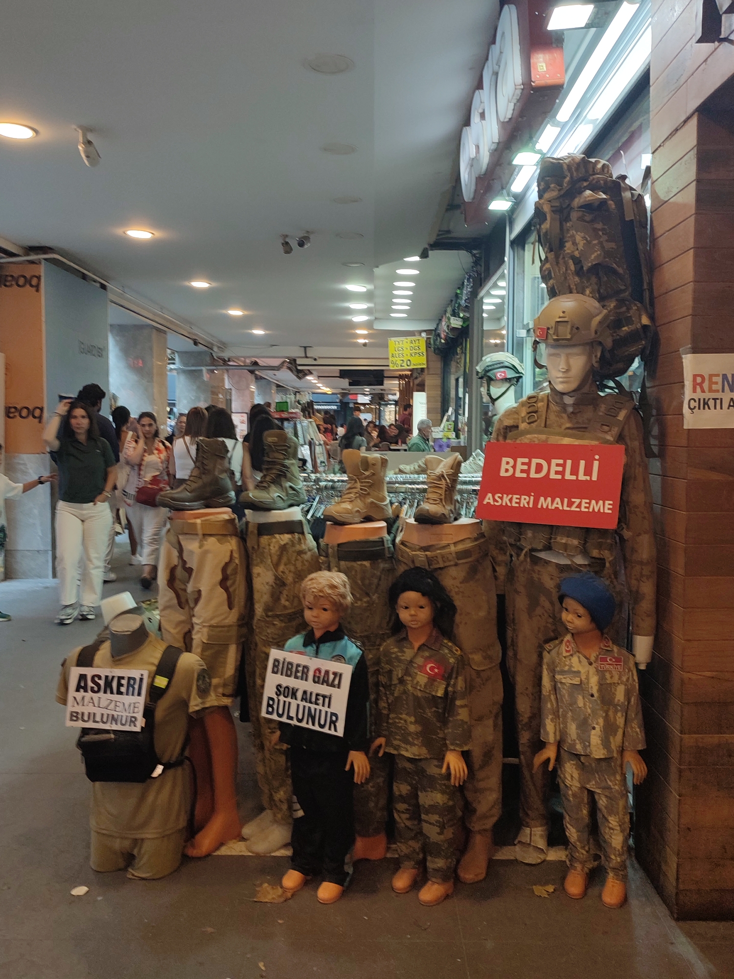 In Kadıköy, Turkey, a street-level view captures a storefront display featuring mannequins dressed in military-style clothing and gear. Several mannequins are positioned to showcase camouflage uniforms, boots, and backpacks, with some holding signs. One prominent sign reads "BEDELLI ASKERI MALZEME," indicating military supplies available for purchase, possibly related to conscription deferment services. Another sign reads "ASKERI MALZEME BULUNUR" (Military supplies available). A smaller sign displays "BIBER GAZI SOK ALETI BULUNUR" (Pepper gas, stun gun available). Pedestrians walk in the background along the covered walkway. The environment appears to be a bustling market or shopping street.
