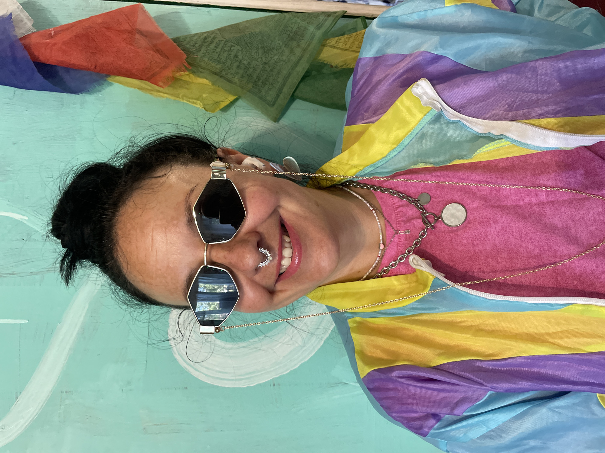The image shows a close-up of a woman in Saint Petersburg, Russia, against a teal wall with abstract white paint strokes. She is wearing large, angular, silver-rimmed sunglasses with a chain attached to one arm. She has a septum piercing with a silver, crescent-shaped jewelry piece. Her hair is pulled up into a messy bun. She is also wearing a bright pink t-shirt, a colorful windbreaker jacket with diagonal panels of purple, yellow, blue, and light green, and several layered necklaces. A string of small white beads with a pink heart charm hangs around her neck, layered with a silver chain that has a circular pendant. In the background, to the right of the woman, prayer flags in various colors, including red, blue, yellow, and green, are attached to the wall. The lighting suggests an indoor setting.