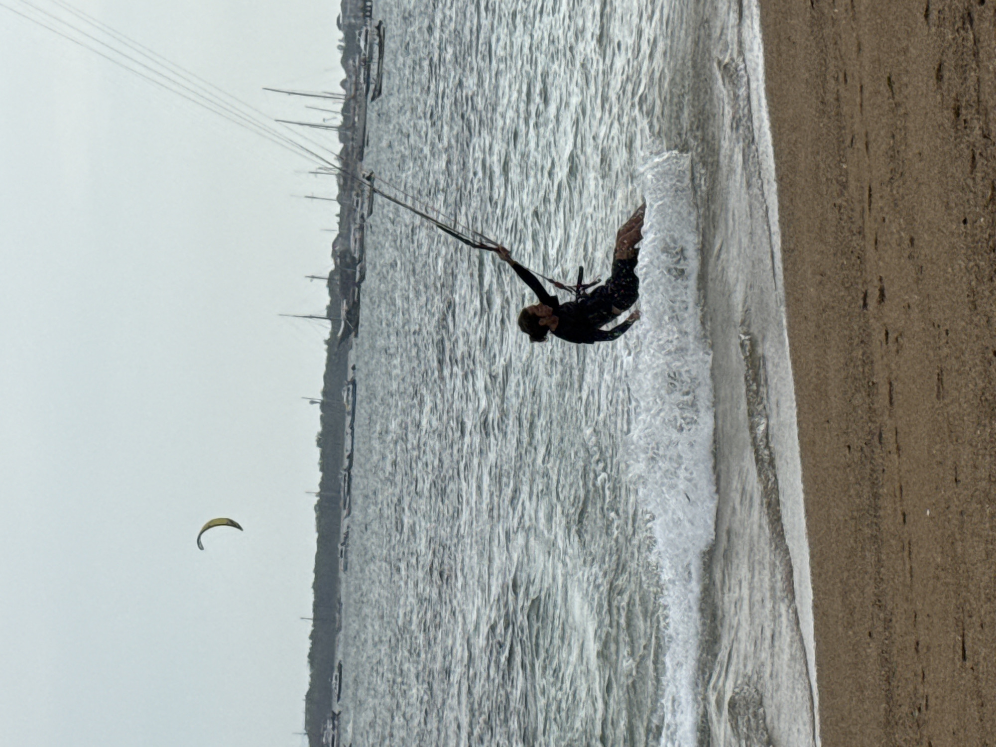 A person is kitesurfing in the ocean off the coast of Denpasar, Indonesia. The kitesurfer, wearing a black wetsuit, is in mid-air, being pulled by a yellow kite. They are kicking up a spray of water as they ride on a wave. In the background, there are several boats anchored in the water and a hazy shoreline. The sky is overcast. The beach in the foreground is made of sand with some footprints visible.