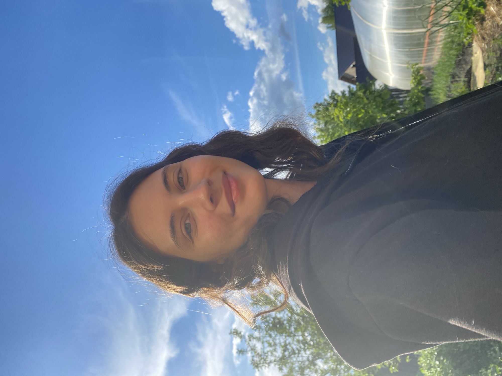 A young woman is shown in a selfie taken outside under a blue sky with scattered clouds. A small, translucent greenhouse is visible in the background along with trees. She is wearing a black hoodie. The location cannot be determined.