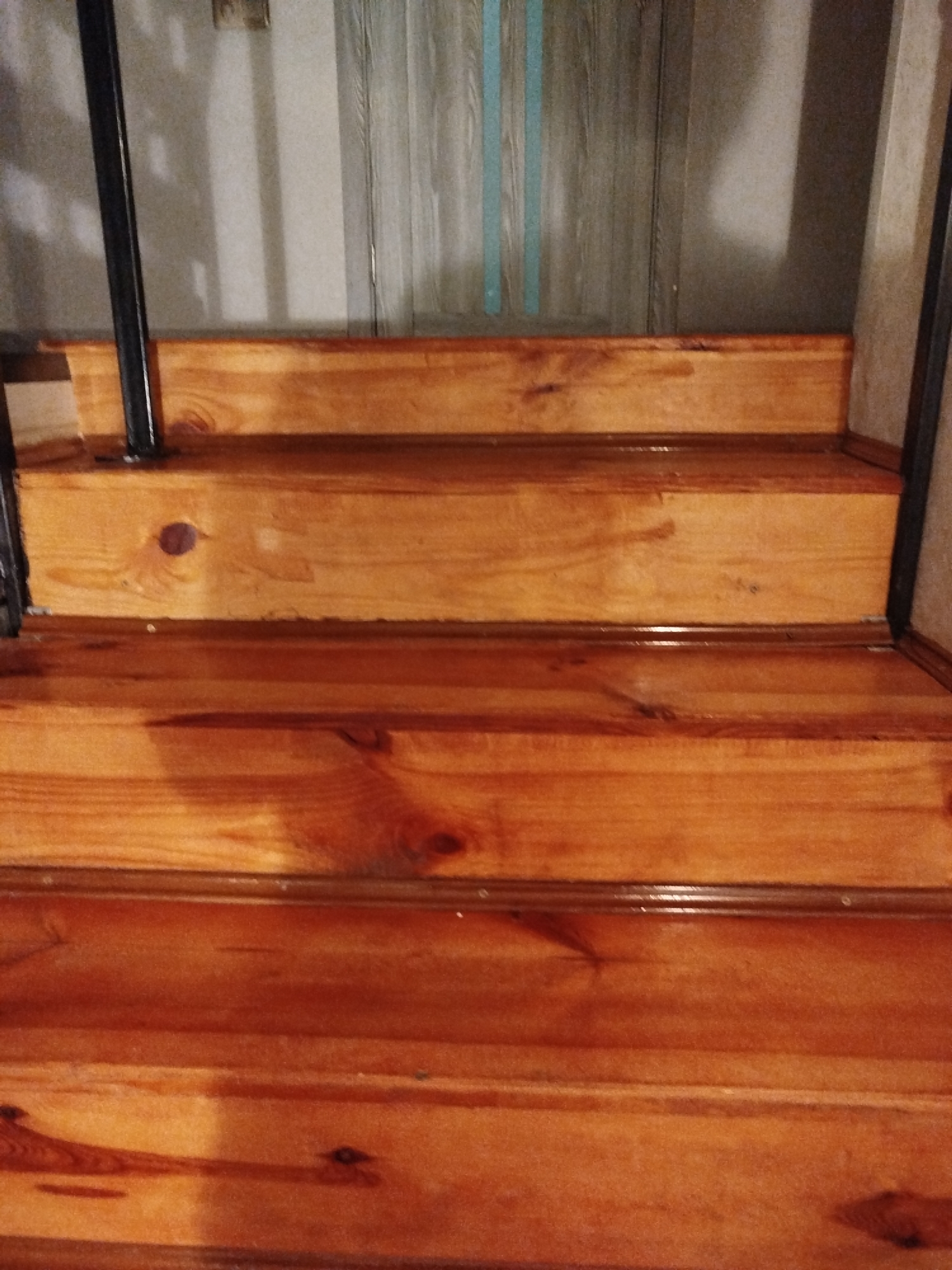 The image shows a close-up view of wooden stairs. The stairs are made of light-colored wood with visible grain and knots, and they appear to be varnished. The risers and treads of the stairs are clearly defined. There is a black metal railing visible on the left side of the image. In the background, there is a wall with a vertical line pattern, possibly wallpaper or a decorative element, and a faint shadow cast on the wall. The lighting suggests an indoor environment, likely a home. The location in Крывасельскі сельскі Савет, Belarus is not directly identifiable from the visual information in the image.