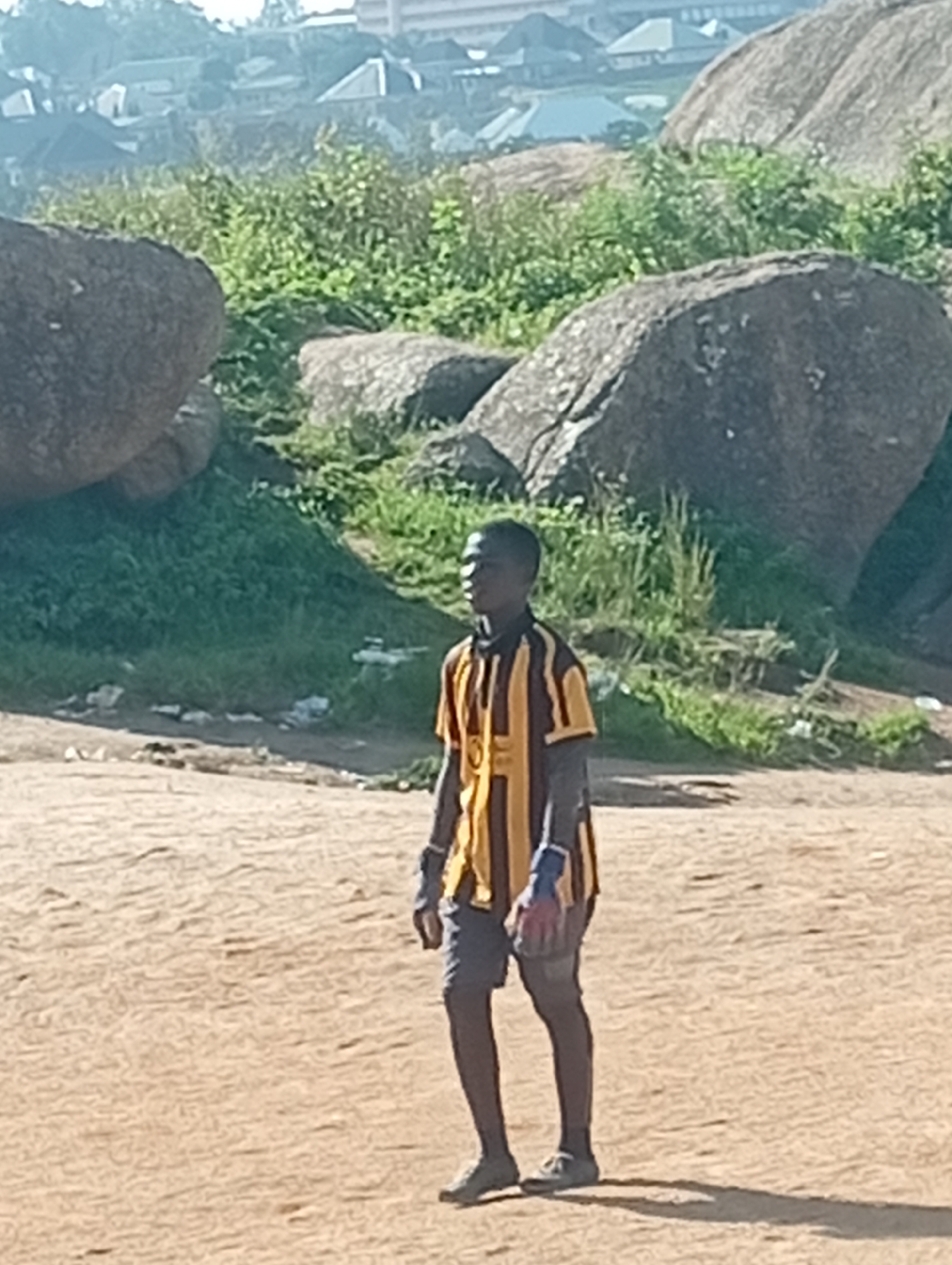 A young Black male, wearing a striped yellow and brown long-sleeved shirt and dark shorts, stands on a dirt field. He has a goalkeeper's glove on his left hand. In the background, there are large rocks, green grass, and a distant view of buildings and trees. The lighting suggests it is daytime.