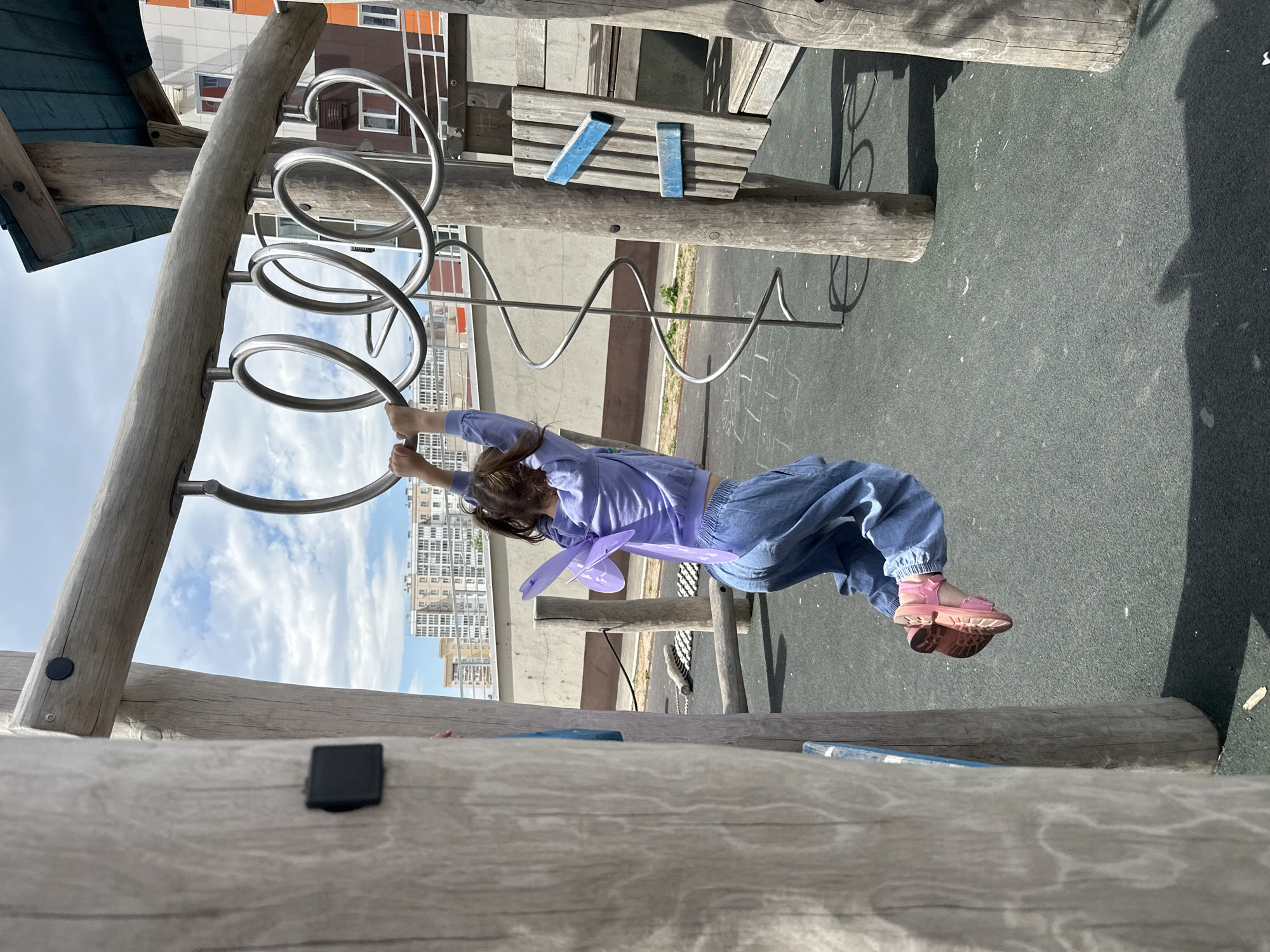 A young girl is hanging from a spiral metal climbing structure in a playground. She is wearing a blue jacket and pants, and pink sandals. The playground is located in Khabarovsk, Russia. In the background, there are residential buildings. The image is rotated, with the sky and buildings appearing to be on their side.