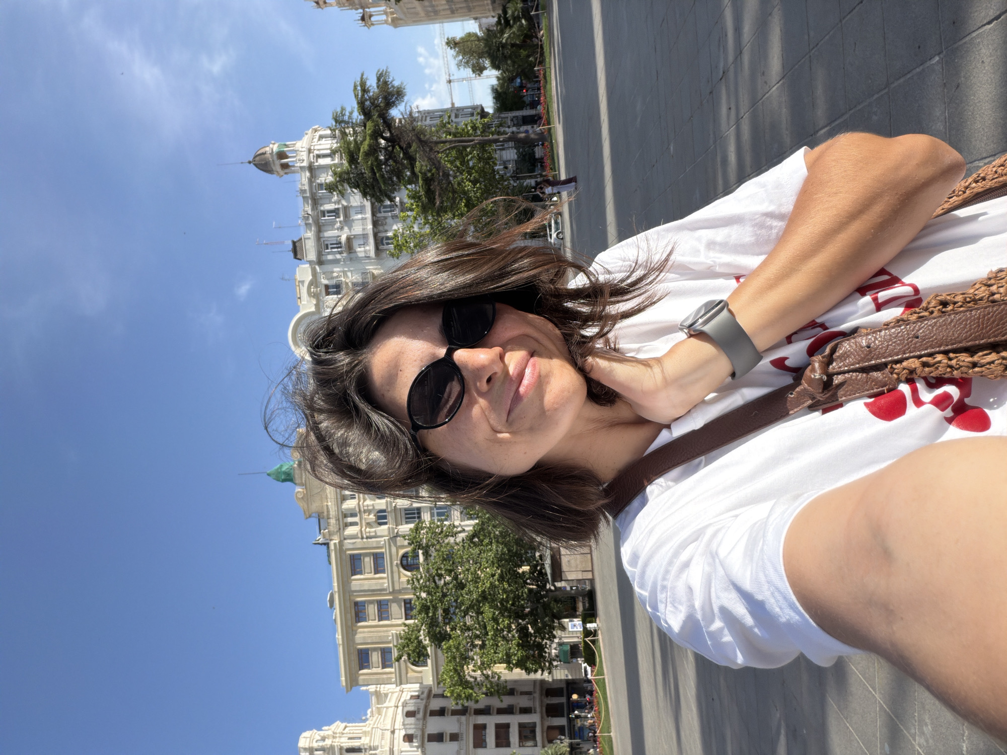 A woman is taking a selfie in front of a large, ornate building in the city of València, Spain. The woman has dark hair and is wearing sunglasses and a white t-shirt with red lettering. She has her hand on her chin and is smiling. In the background, the building is light-colored with many windows and architectural details. There are trees and a blue sky with a few faint clouds. The ground in front of the building is paved with stone.