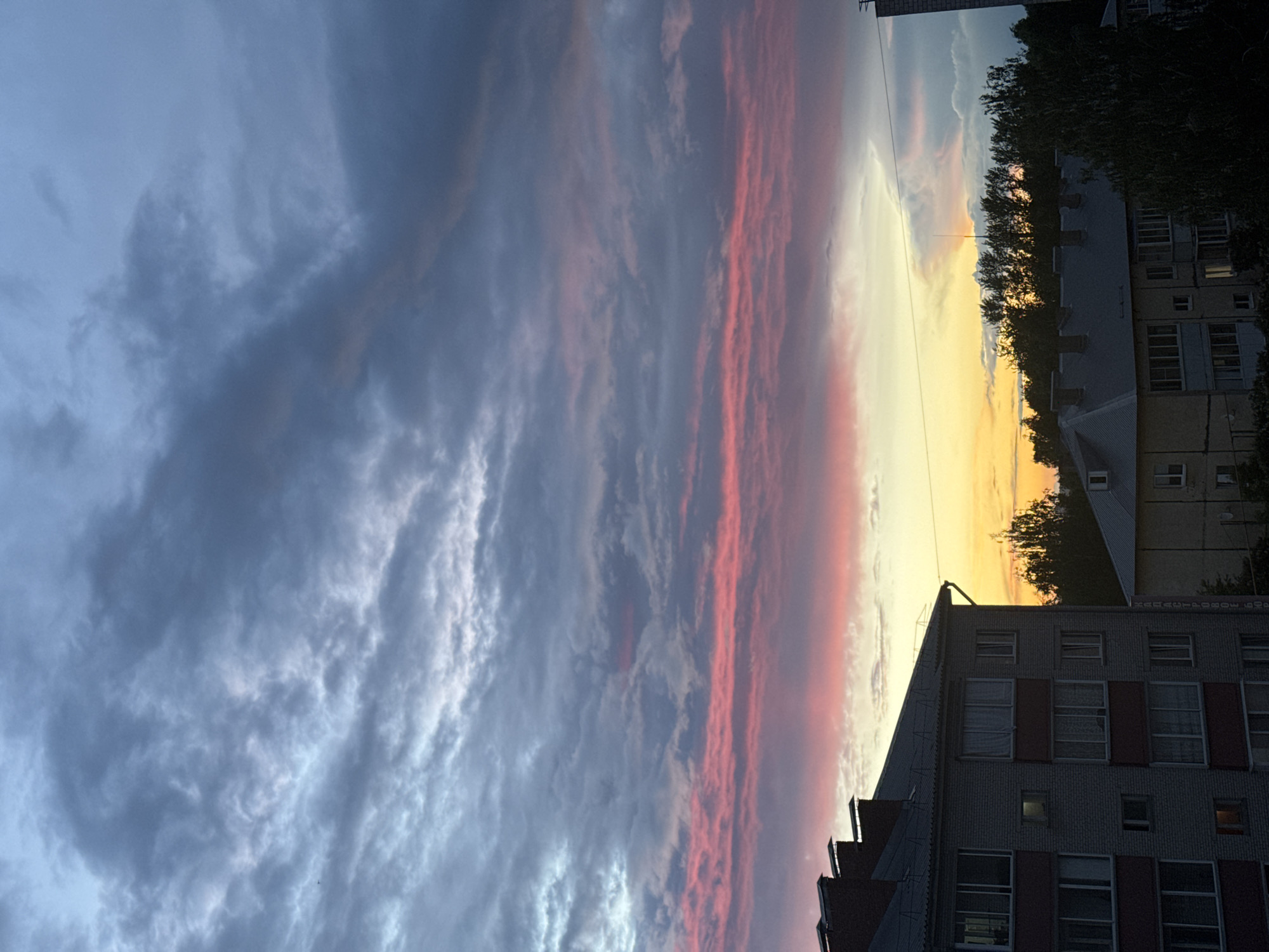 The image shows a sunset sky in what appears to be Zav'yalovo, Russia. The sky displays a gradient from a dark blue-gray cloud cover on the left to a band of pink and orange hues closer to the horizon on the right. The clouds are layered and textured.
In the lower right of the frame, the corner of a multi-story building is visible. The building is made of brick and has multiple windows. Behind the building are trees and possibly more buildings in the distance.