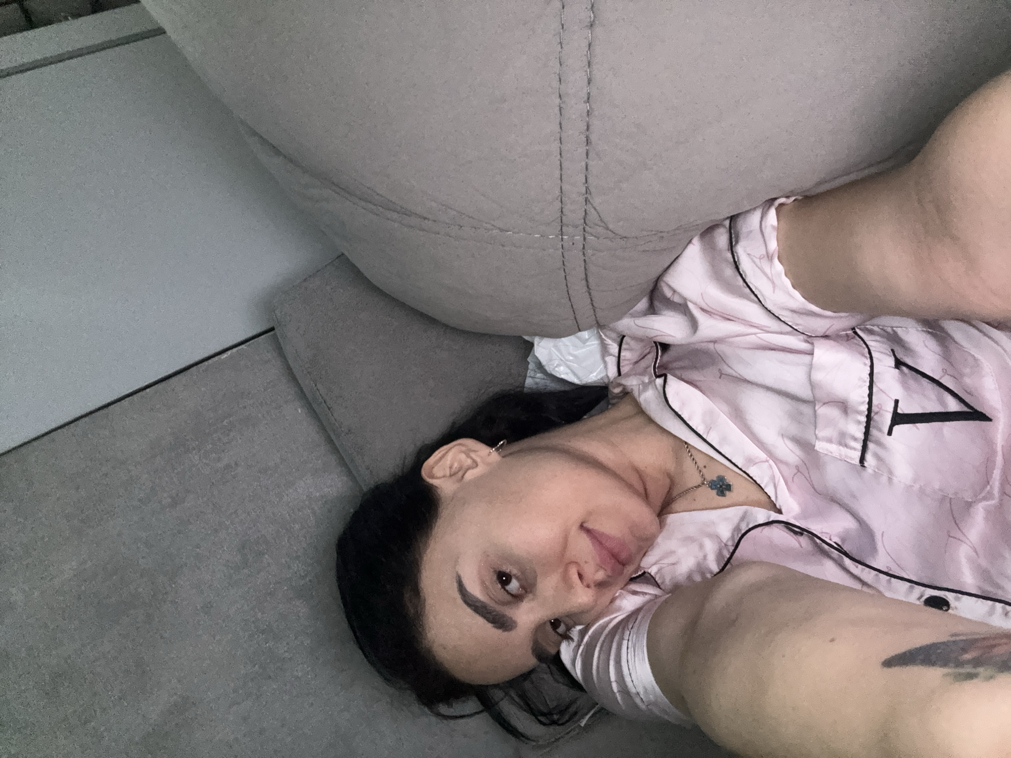 A woman is lying on a gray couch, wearing a light pink pajama top with black piping and a large black "1" on the right breast. She has dark hair, fair skin, and a visible tattoo on her upper arm. She is positioned at an angle, with her head resting on the couch and her body partially supported by a large, gray cushion or pillow. Part of a white wall is visible in the background to the left. The location is likely indoors and could be in Tbilisi, Georgia, but there are no landmarks to confirm the location.
