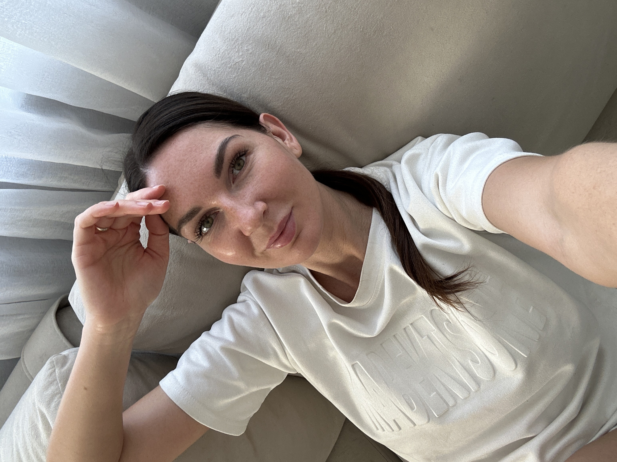 A young woman with brown hair and green eyes is lying on a beige couch, looking at the camera. She is wearing a white t-shirt with embossed text. Her left arm is bent, with her hand resting on her forehead, and she is wearing a ring on her left ring finger. Her right arm is bent and held in front of her. She has makeup on, including mascara and lip gloss. The background consists of beige couch cushions and sheer white curtains. The image appears to be a selfie, taken indoors in what looks like a residential setting. There is no visible indication of the city of Chaikovsky, Russia.