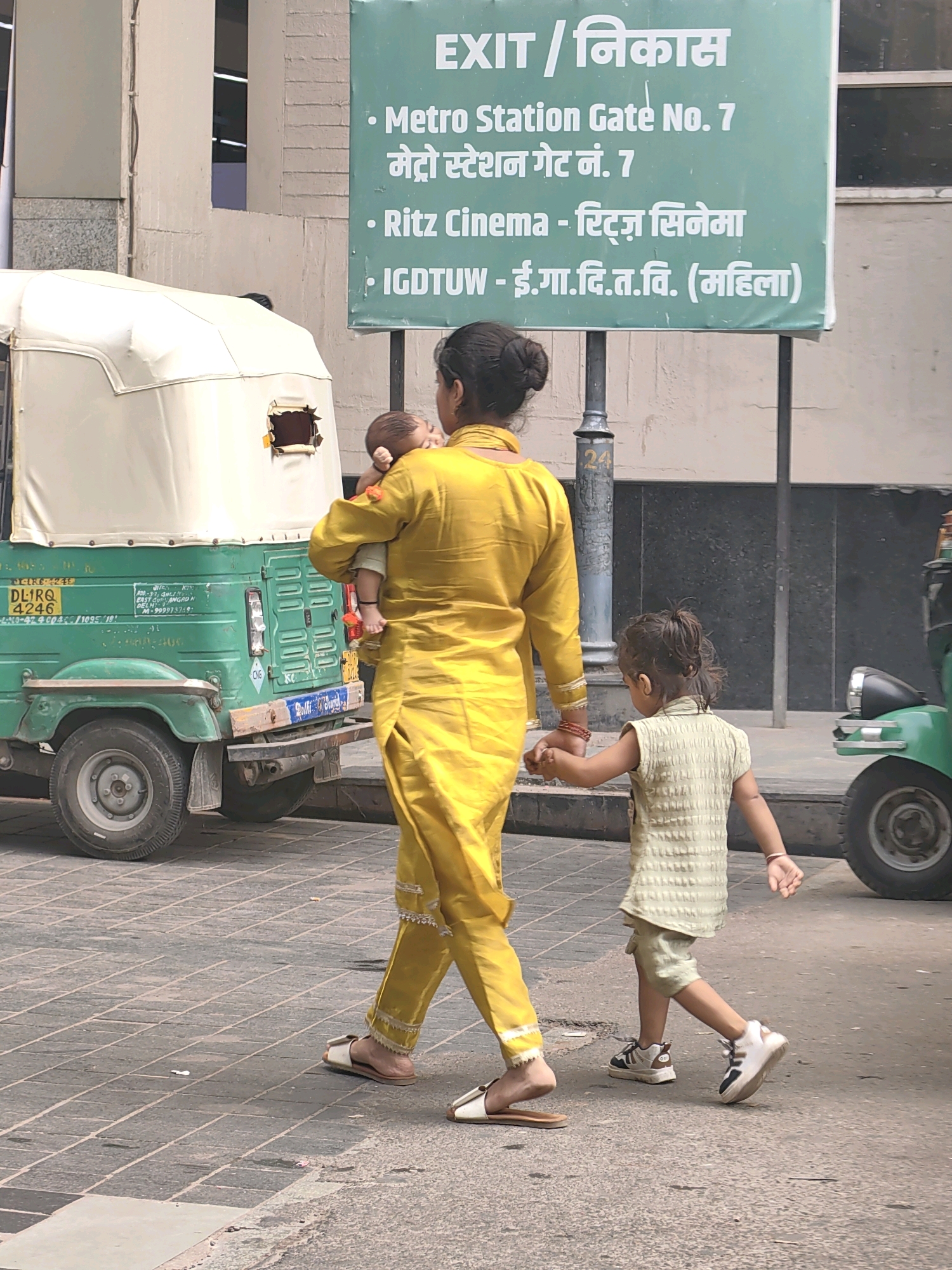The image depicts a street scene in Delhi, India, likely near a metro station, as indicated by the sign in the background. A woman in a vibrant yellow salwar kameez is walking, carrying a baby. Beside her, a young girl in a light green outfit is holding her hand. They are walking on a paved sidewalk, with a green auto-rickshaw visible to their left. The auto-rickshaw has license plates and various markings, suggesting it is a commercial vehicle. In the background, a large green sign with white text announces "EXIT / विकास" and lists directions to "Metro Station Gate No. 7," "Ritz Cinema," and "IGDTUW - ई.गा.दि.त.वि. (महिला)." The environment appears to be an urban setting with buildings and street infrastructure. The lighting suggests it is daytime, possibly under cloudy or hazy conditions.