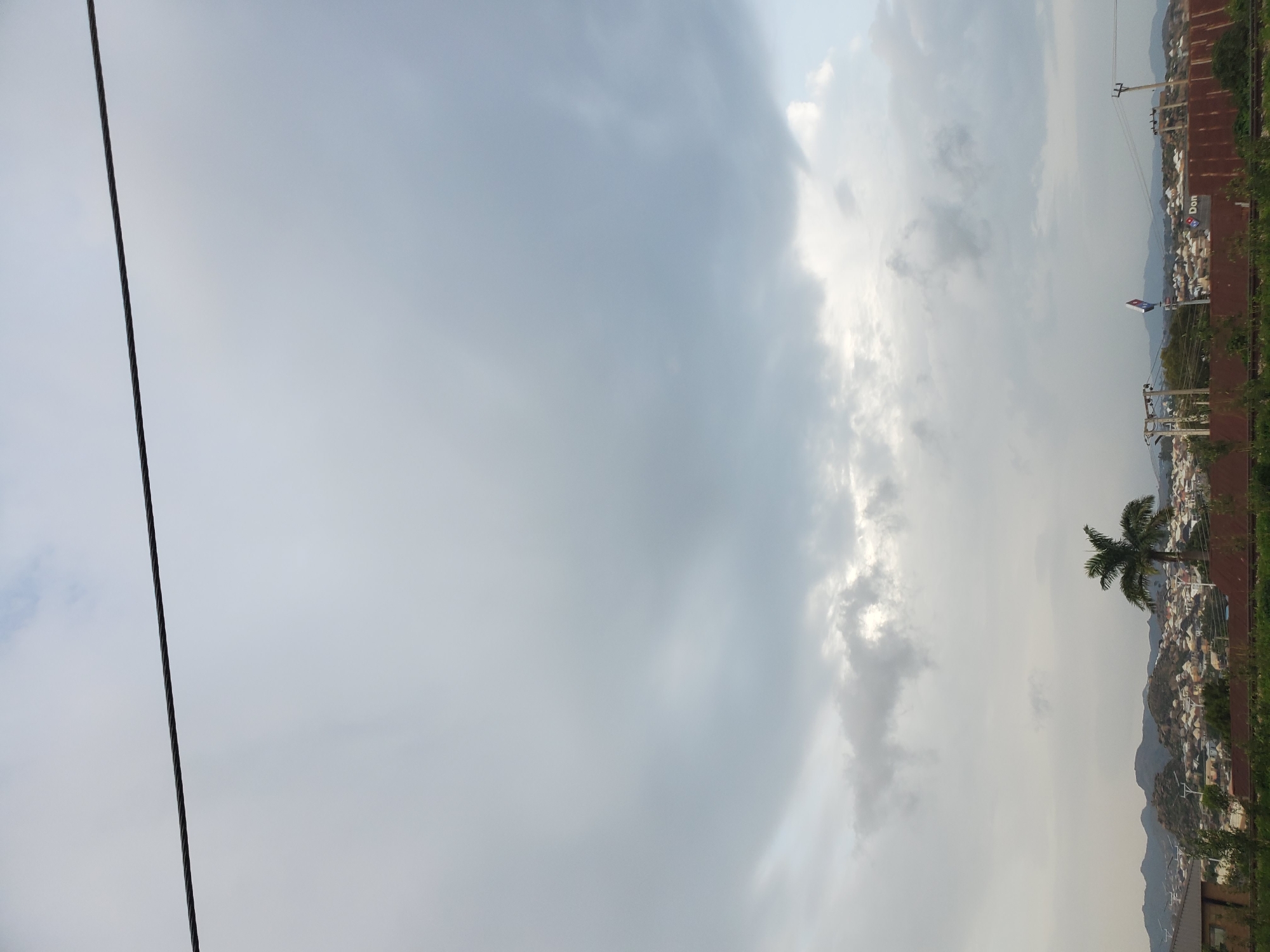 The image depicts a panoramic view of a residential area in Jos, Nigeria, under a cloudy sky. A thick dark cable stretches horizontally across the upper portion of the frame, dividing the sky. In the middle ground, a solitary palm tree stands prominently. Beyond the tree, a dense settlement of houses with red roofs is nestled against a backdrop of rocky hills. Power lines and utility poles are visible throughout the scene, indicating infrastructure development. Two Domino's Pizza signs are discernible on a building in the distance, suggesting commercial activity within the residential area. The overall atmosphere is overcast, with diffused light, hinting at potentially impending rain.